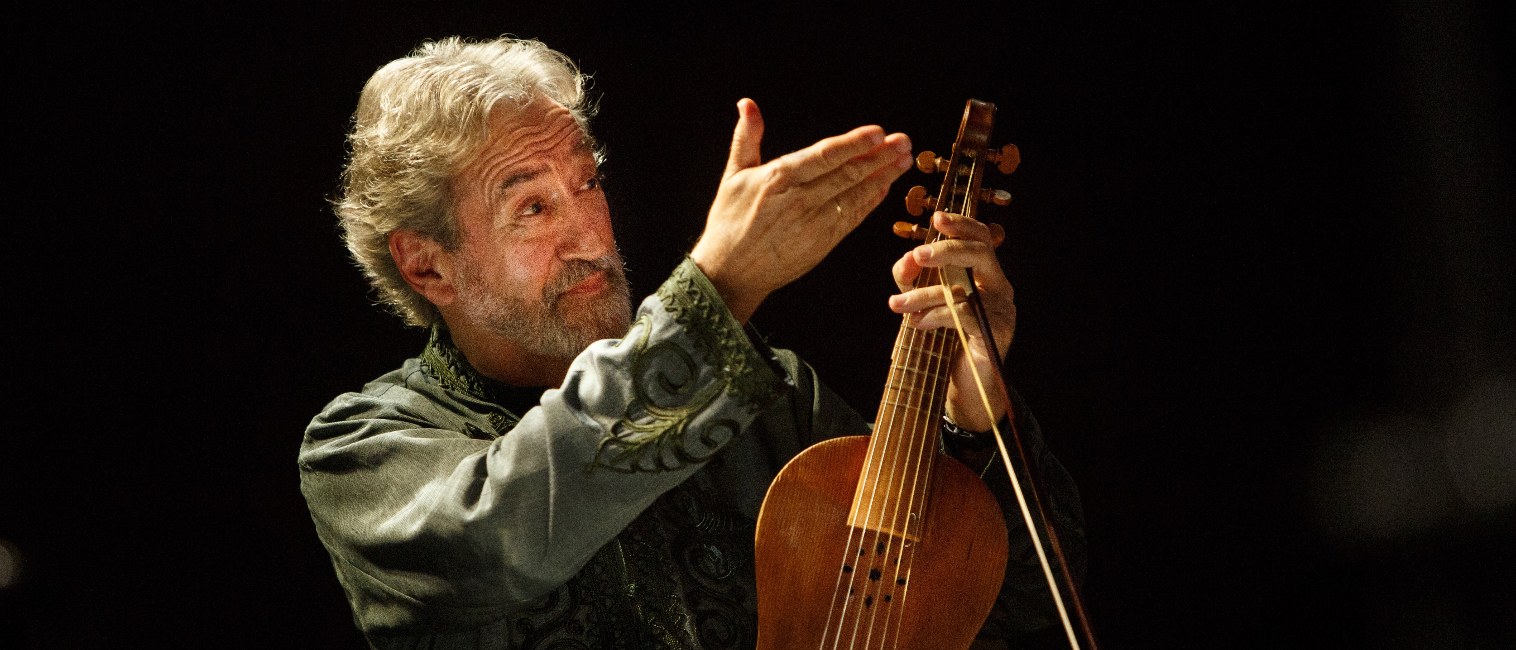 Jordi Savall, with grey hair and a green patterned shirt, holds a wooden stringed instrument in his hand. He is standing on a dimly lit stage. Sheet music and a music stand can be seen in the foreground.