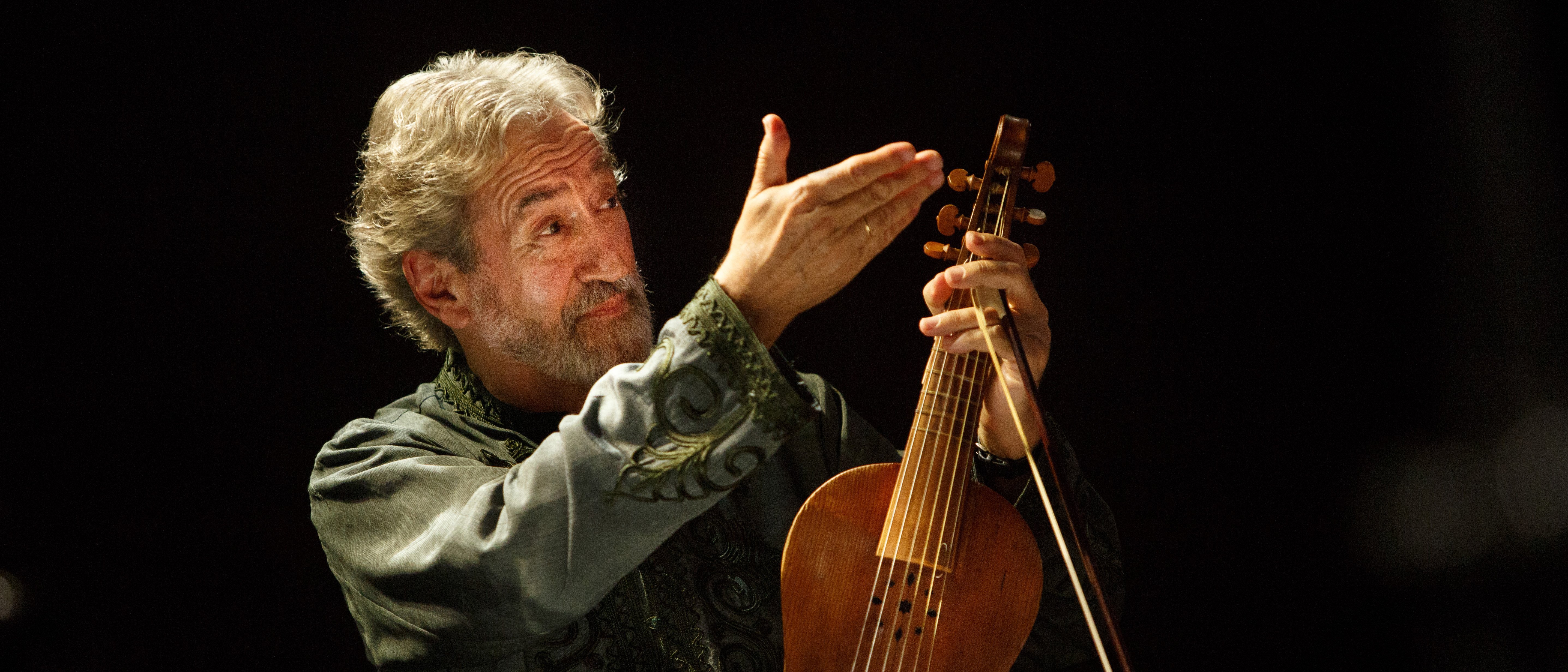Jordi Savall, mit grauem Haar und einem grün gemusterten Hemd, hält ein hölzernes Saiteninstrument in der Hand. Er steht auf einer schwach beleuchteten Bühne. Im Vordergrund sind Notenblätter und ein Notenpult zu sehen.