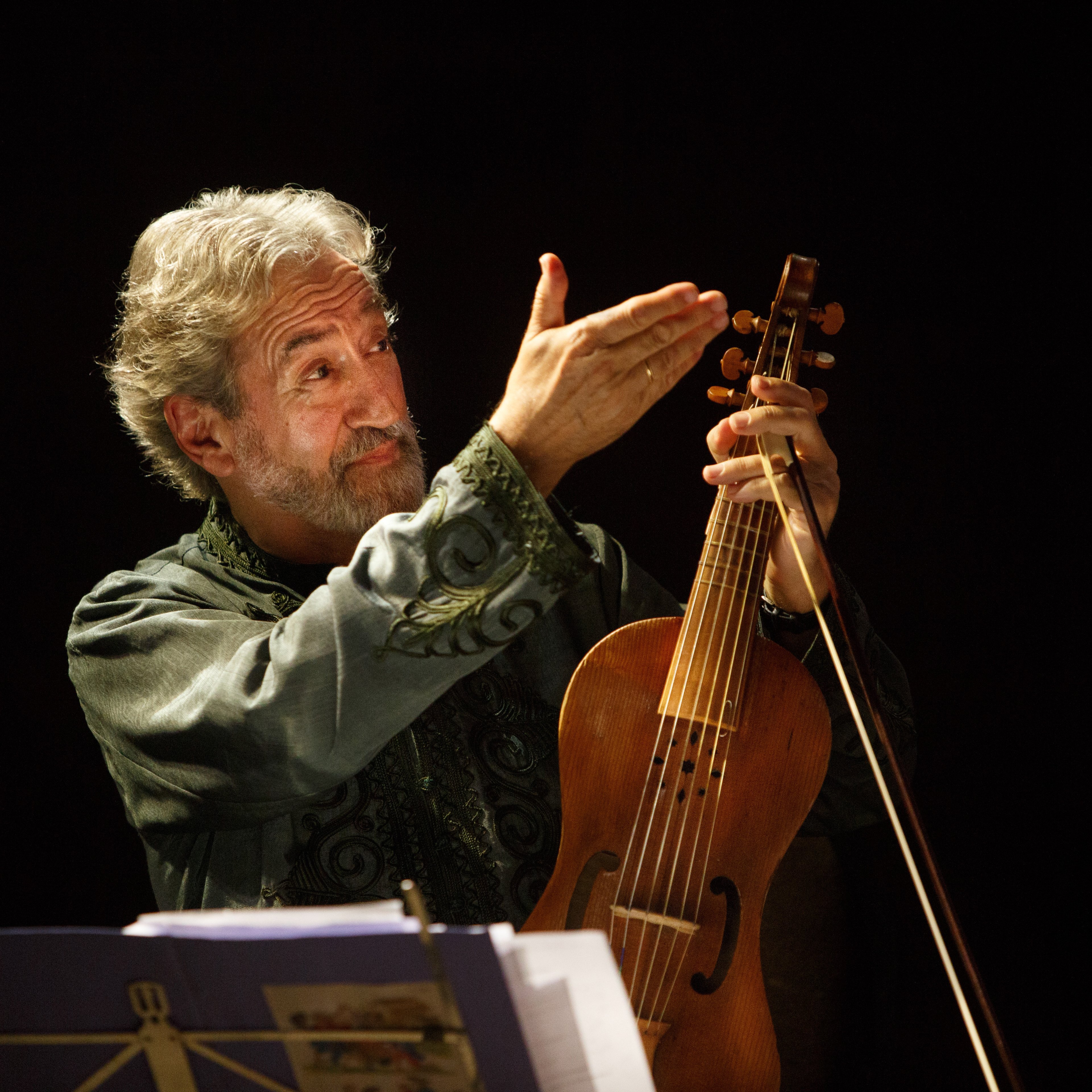 Jordi Savall, mit grauem Haar und einem grün gemusterten Hemd, hält ein hölzernes Saiteninstrument in der Hand. Er steht auf einer schwach beleuchteten Bühne. Im Vordergrund sind Notenblätter und ein Notenpult zu sehen.