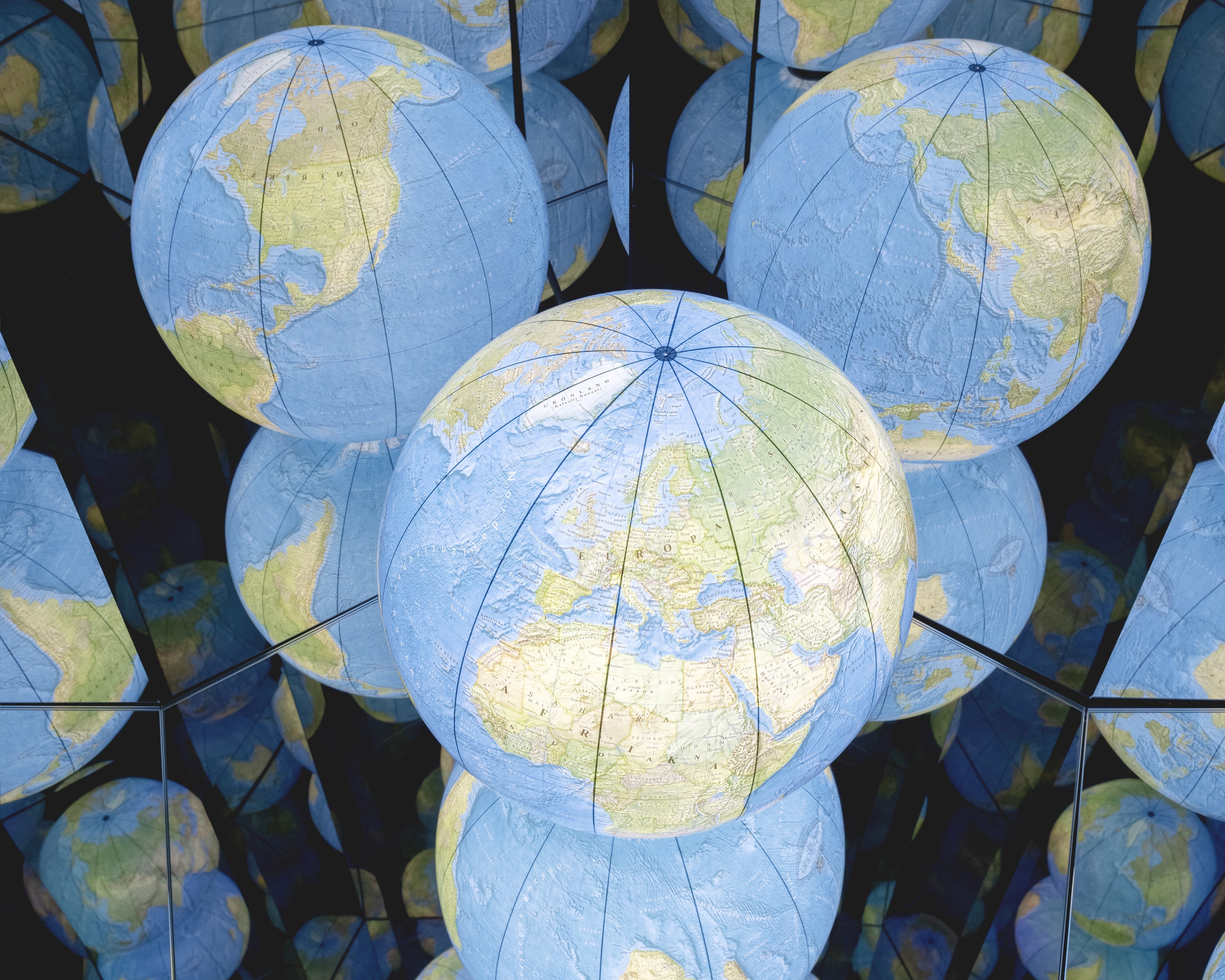 Mehrere blaue Globen mit Weltkarten werden in mehreren Spiegeln reflektiert, wodurch die Illusion endloser Reihen von Globen entsteht. Das Bild hat eine geometrische und symmetrische Komposition.