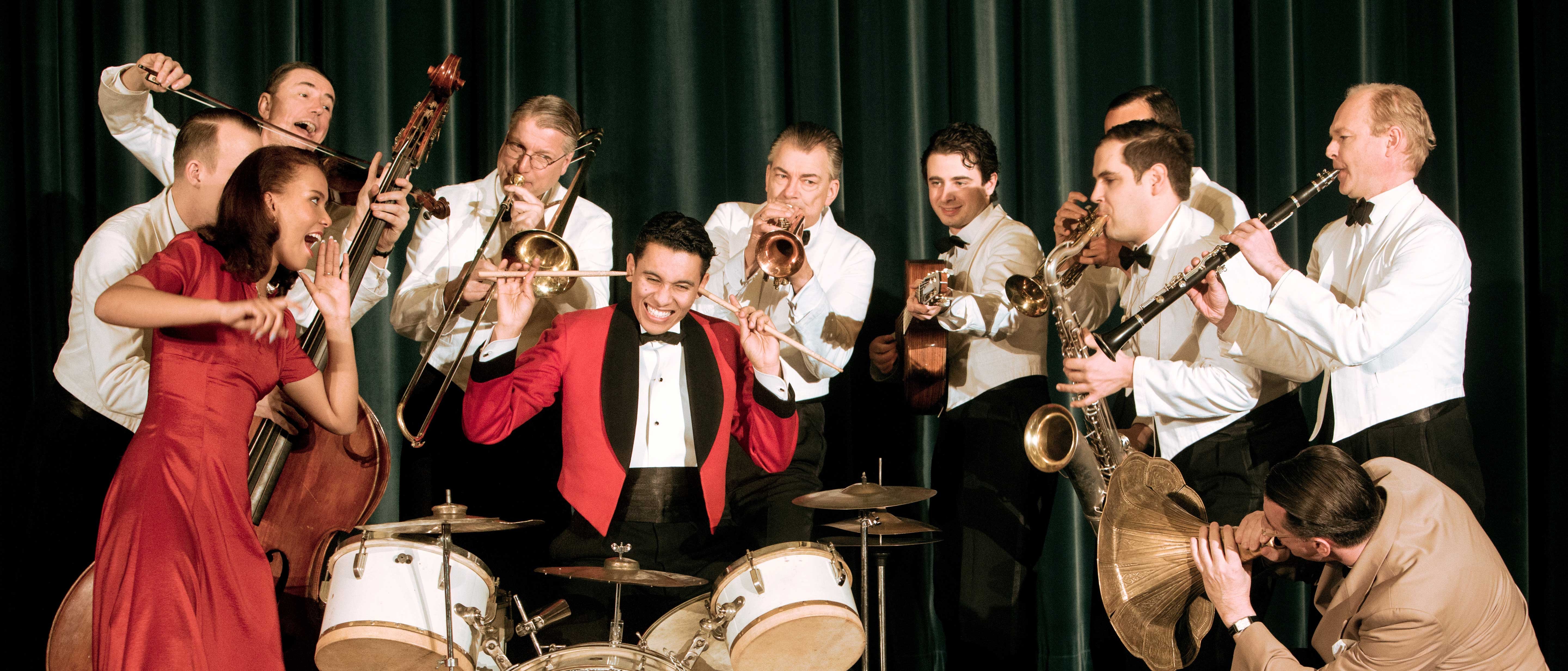 Eine Jazzband in Retro-Kleidung tritt auf der Bühne auf. Der Schlagzeuger in einer roten Jacke lächelt, umgeben von Musikern mit Blechblas-, Streich- und Holzblasinstrumenten. Eine Frau in einem roten Kleid singt fröhlich.