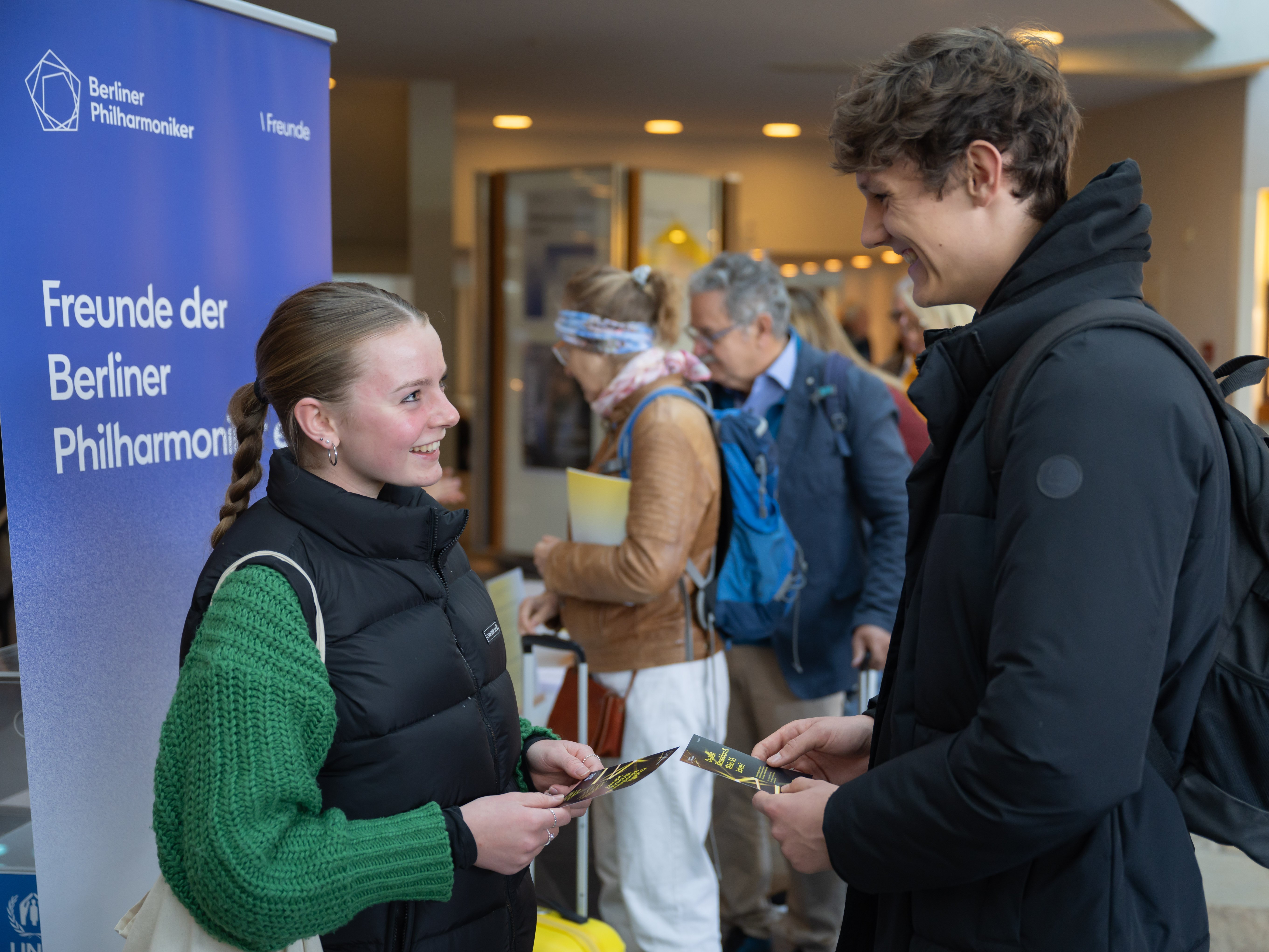 Several people are standing in the foyer of the Berlin Philharmonie, chatting. Some are holding programme booklets. The background shows other guests and architectural details of the foyer.