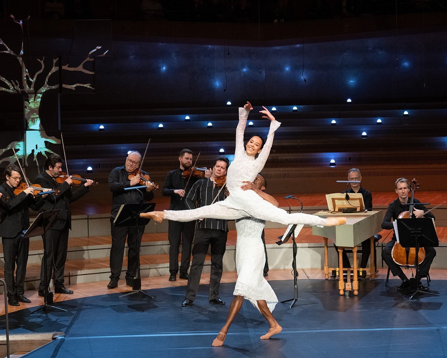 Eine Ballerina in einem weißen Outfit springt anmutig, während ein männlicher Tänzer sie hochhebt; im Hintergrund spielen Musiker auf einer Konzertbühne mit einer beleuchteten, baumähnlichen Requisite Streichinstrumente.