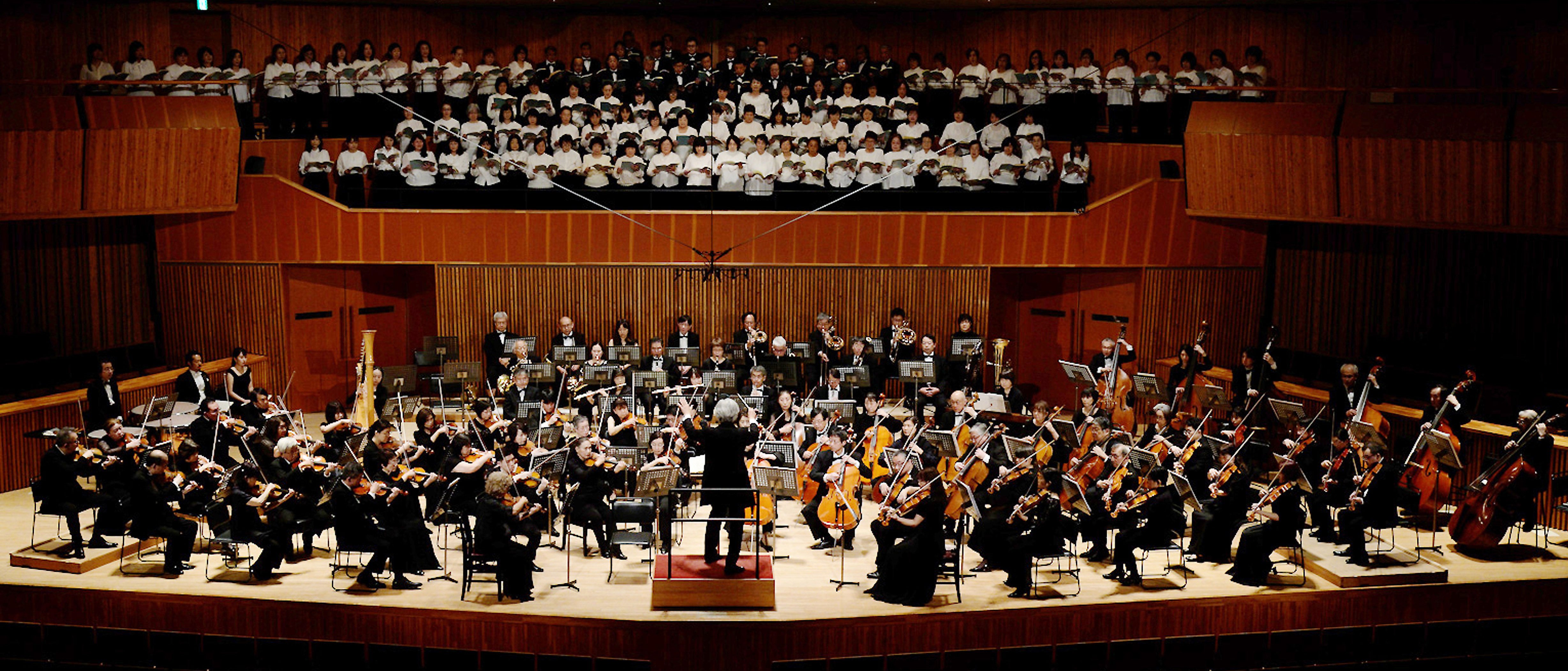 Ein Orchester spielt auf der Bühne eines Konzertsaals unter der Leitung eines Dirigenten. Hinter ihm steht ein großer Chor in weißer Kleidung. Der Saal ist warm beleuchtet, und die Zuschauerplätze sind leer.