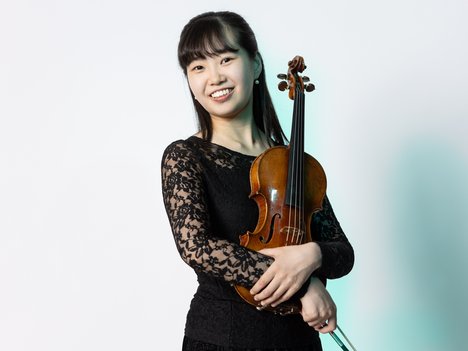 A young woman in a black lace dress smiles while holding a violin and bow across her chest, standing against a plain white background.