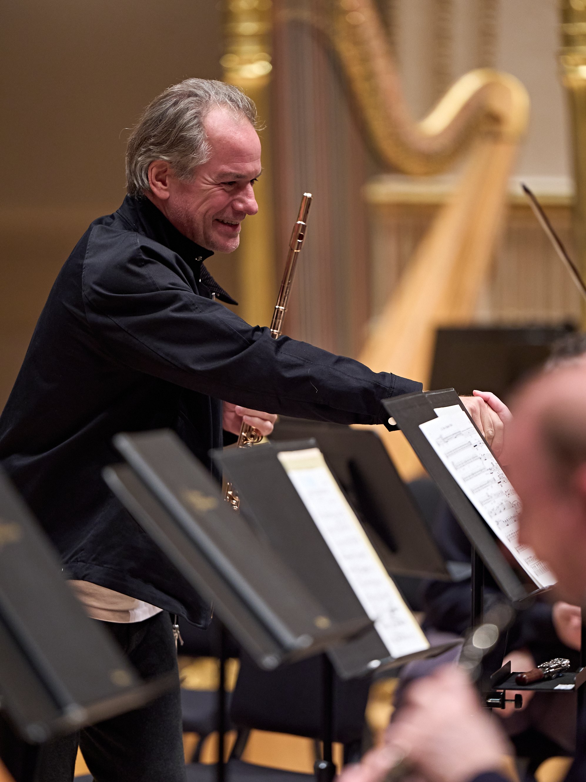 Ein lächelnder Flötist in einer schwarzen Jacke steht mit seinem Instrument in der Orchestergrube, im Hintergrund ist eine Harfe zu sehen.