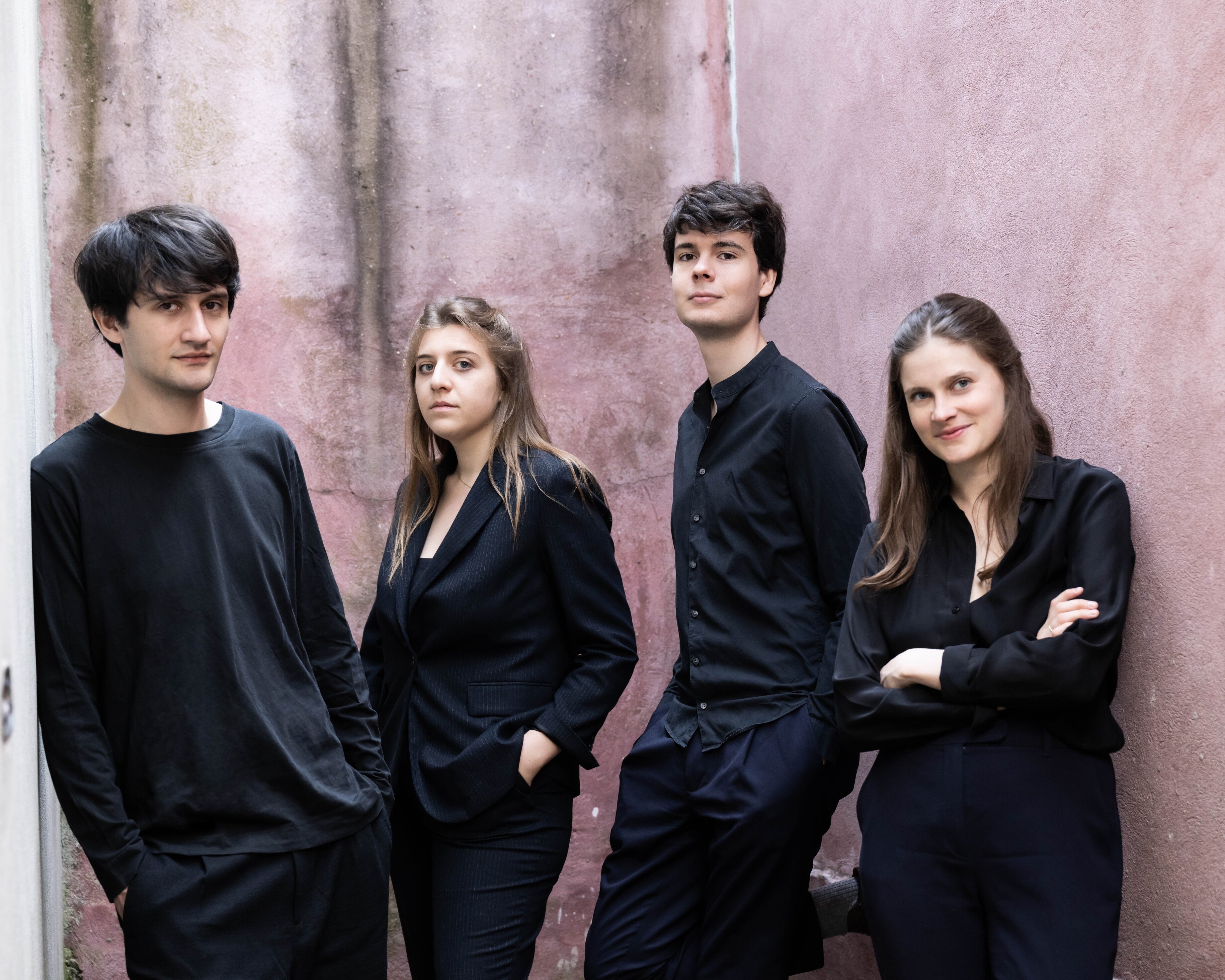 Vier junge Erwachsene stehen in einer engen Gasse mit rosafarbenen Wänden und tragen dunkle, modische Kleidung. Drei schauen in die Kamera, während die Frau rechts mit verschränkten Armen lächelt.