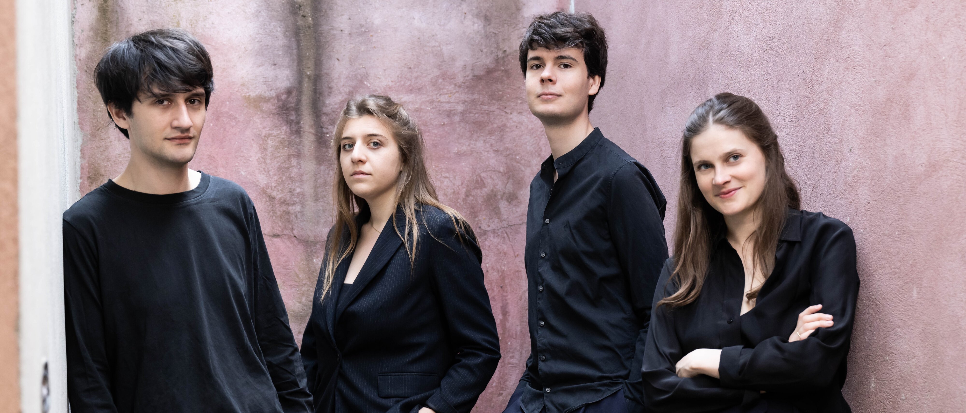 Vier junge Erwachsene stehen in einer engen Gasse mit rosafarbenen Wänden und tragen dunkle, modische Kleidung. Drei schauen in die Kamera, während die Frau rechts mit verschränkten Armen lächelt.
