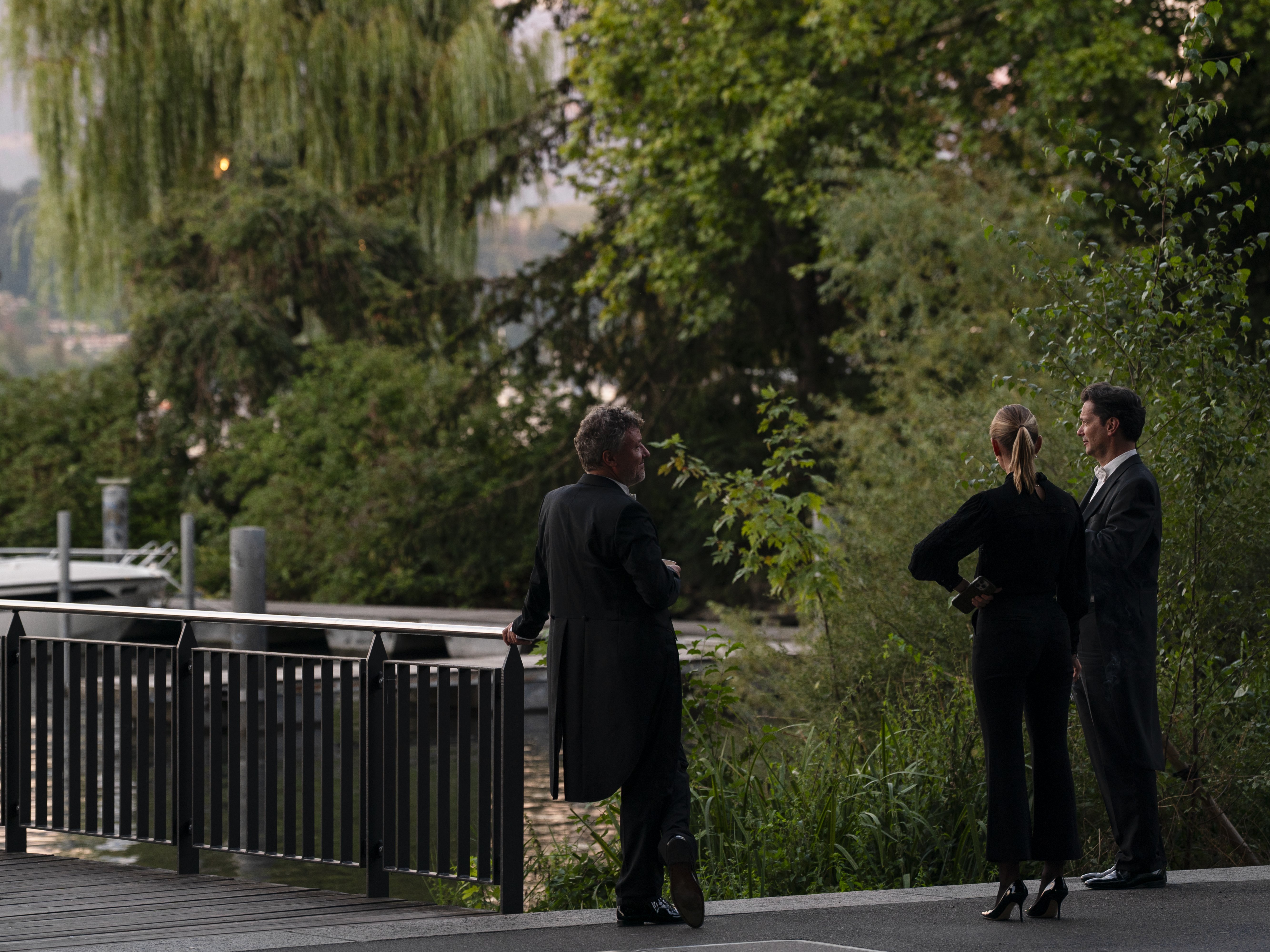 Three people in formal attire stand by a railing near a docked boat, surrounded by lush green trees and foliage, engaged in conversation at dusk.