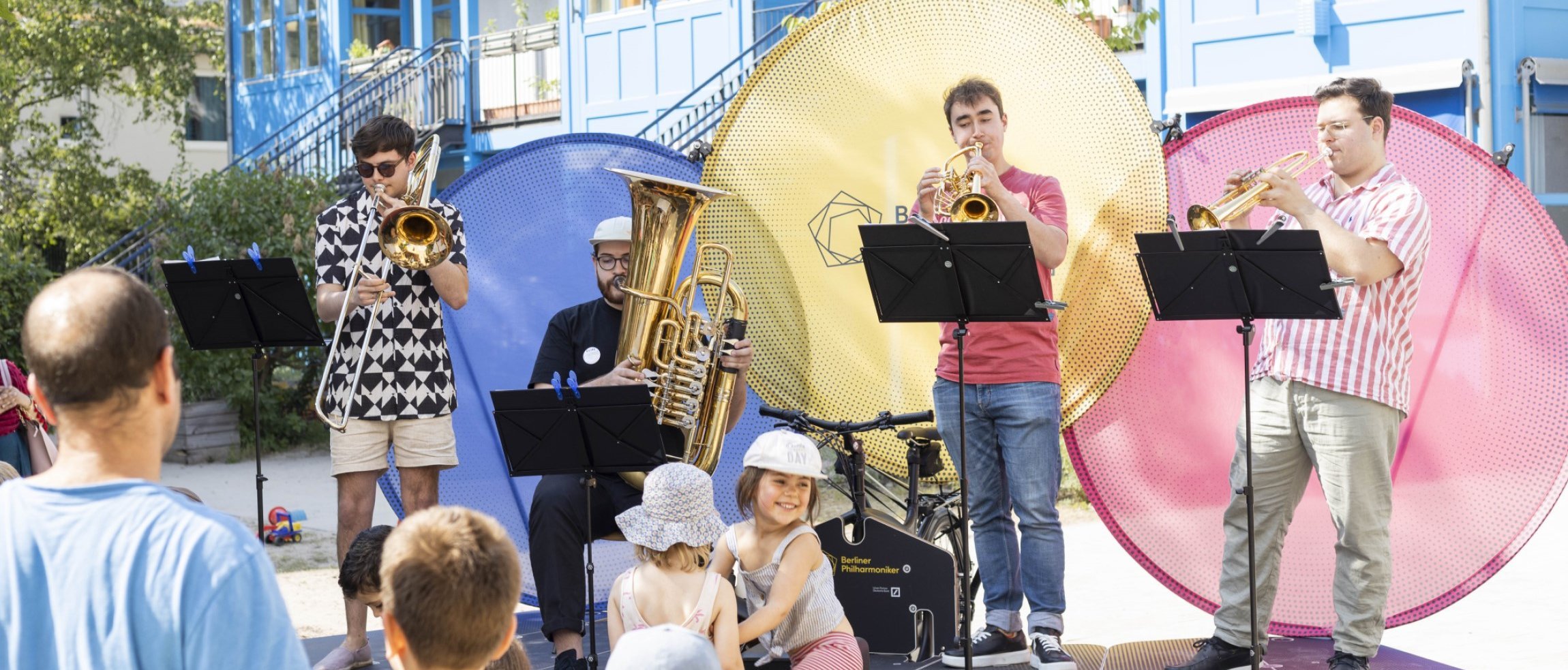 Eine Blaskapelle tritt im Freien vor einem Publikum aus Kindern und Erwachsenen auf. Die Musiker spielen Posaune, Tuba, Trompete und Saxophon vor großen bunten Kreisen als Kulisse.