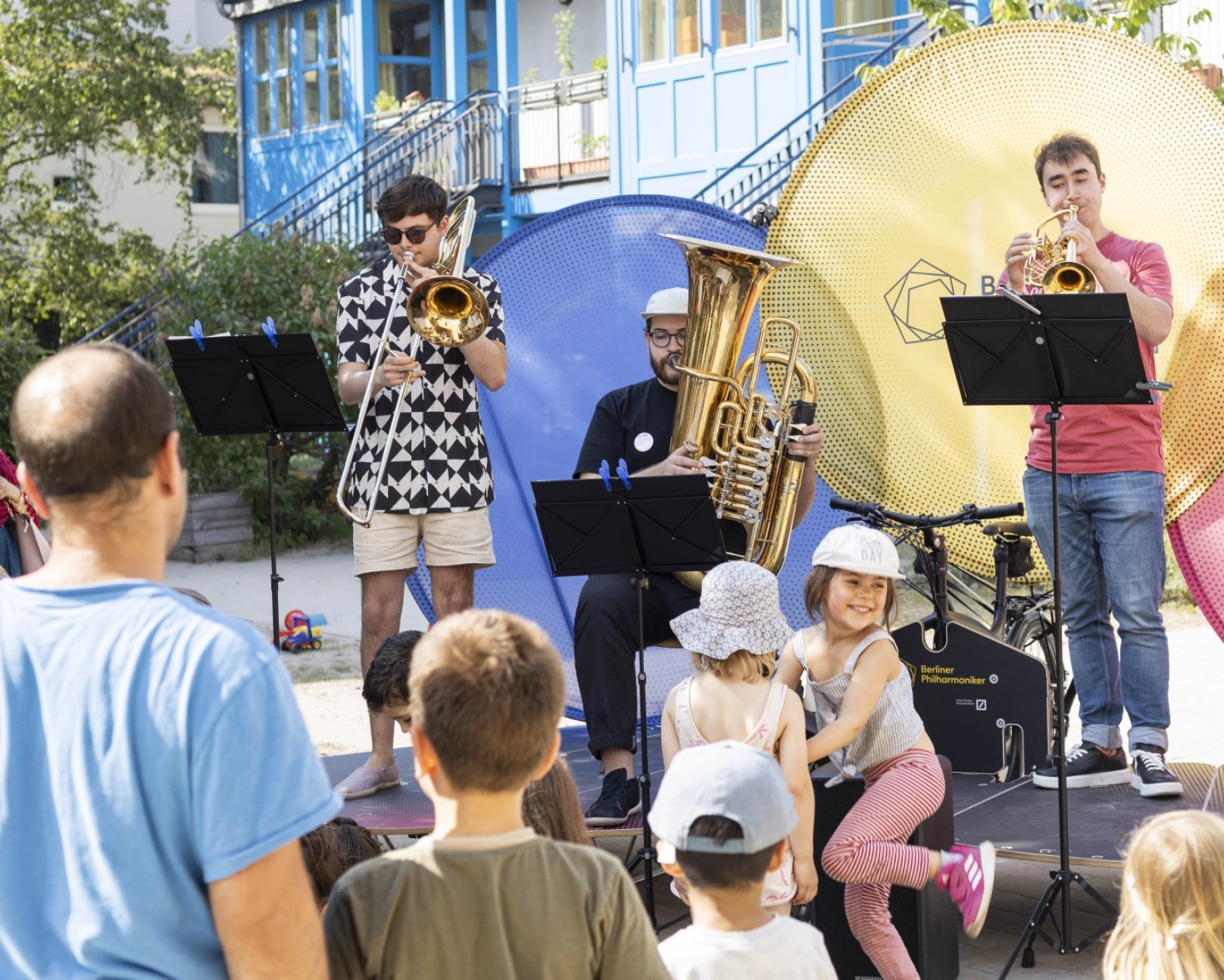 Eine Blaskapelle tritt im Freien vor einem Publikum aus Kindern und Erwachsenen auf. Die Musiker spielen Posaune, Tuba, Trompete und Saxophon vor großen bunten Kreisen als Kulisse.