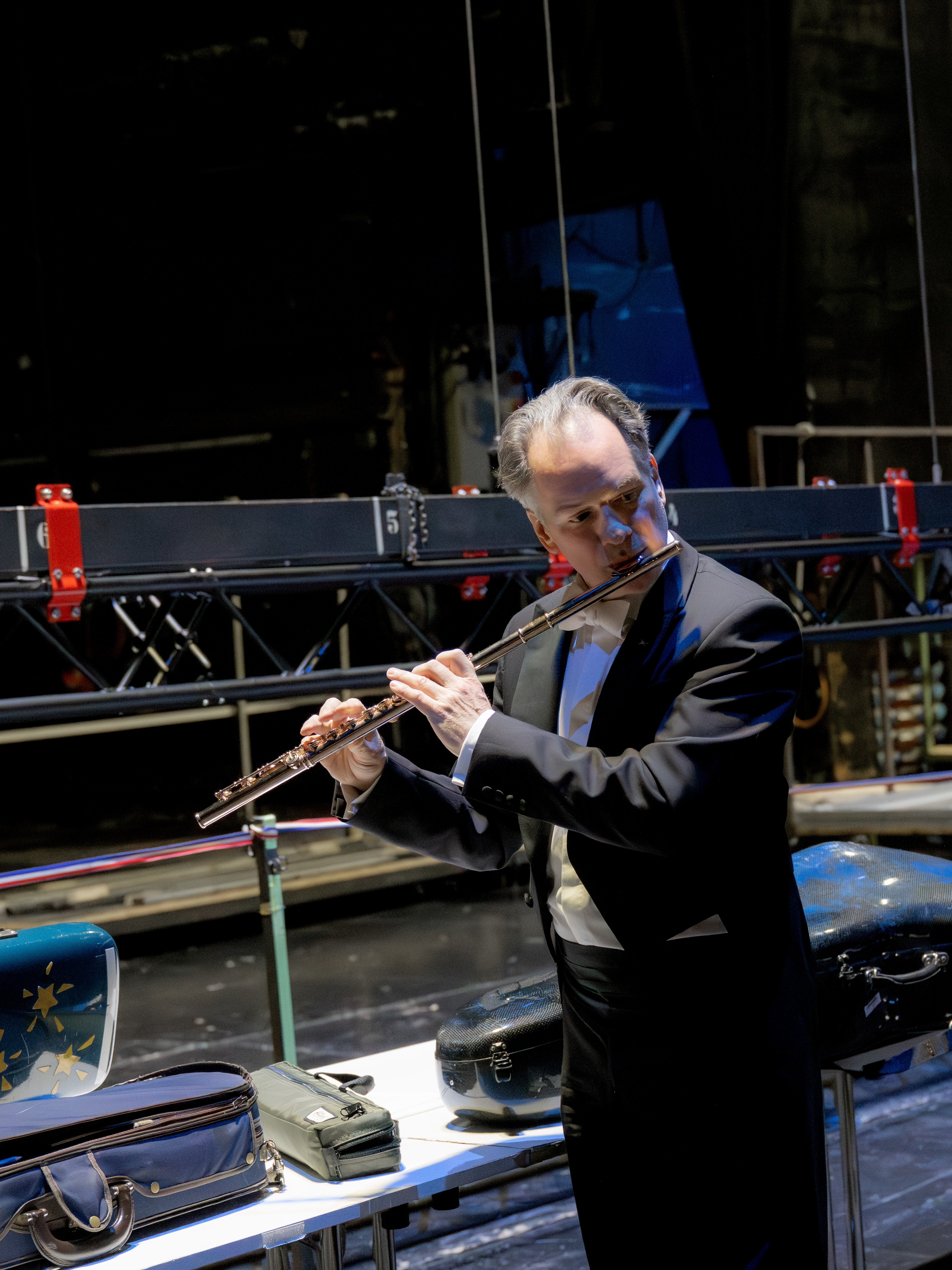 Emmanuel Pahud in formeller Konzertkleidung spielt hinter der Bühne Querflöte und steht neben einem Tisch mit Instrumentenkoffern und Taschen. Im Hintergrund sind Bühnenausstattung und schummrige Beleuchtung zu sehen.