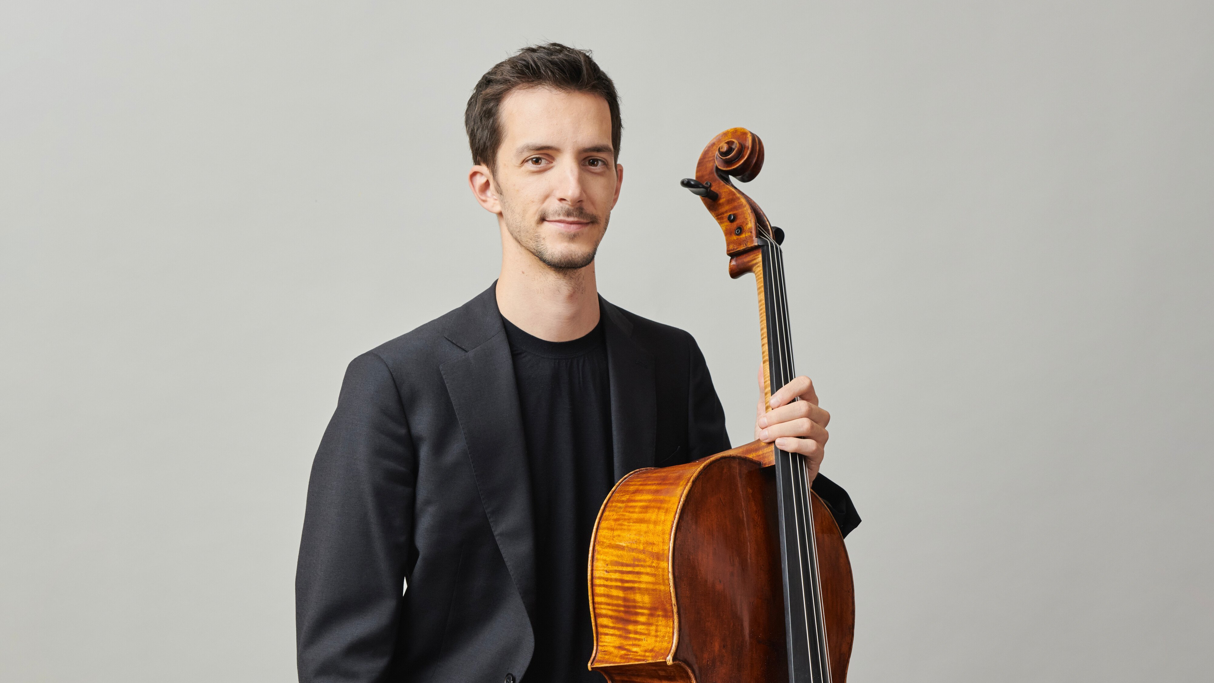 Bruno Delepelaire in schwarzem Anzug und schwarzem Hemd steht mit einem Holzcello in der Hand vor einem schlichten hellgrauen Hintergrund und blickt mit einem leichten Lächeln in die Kamera.
