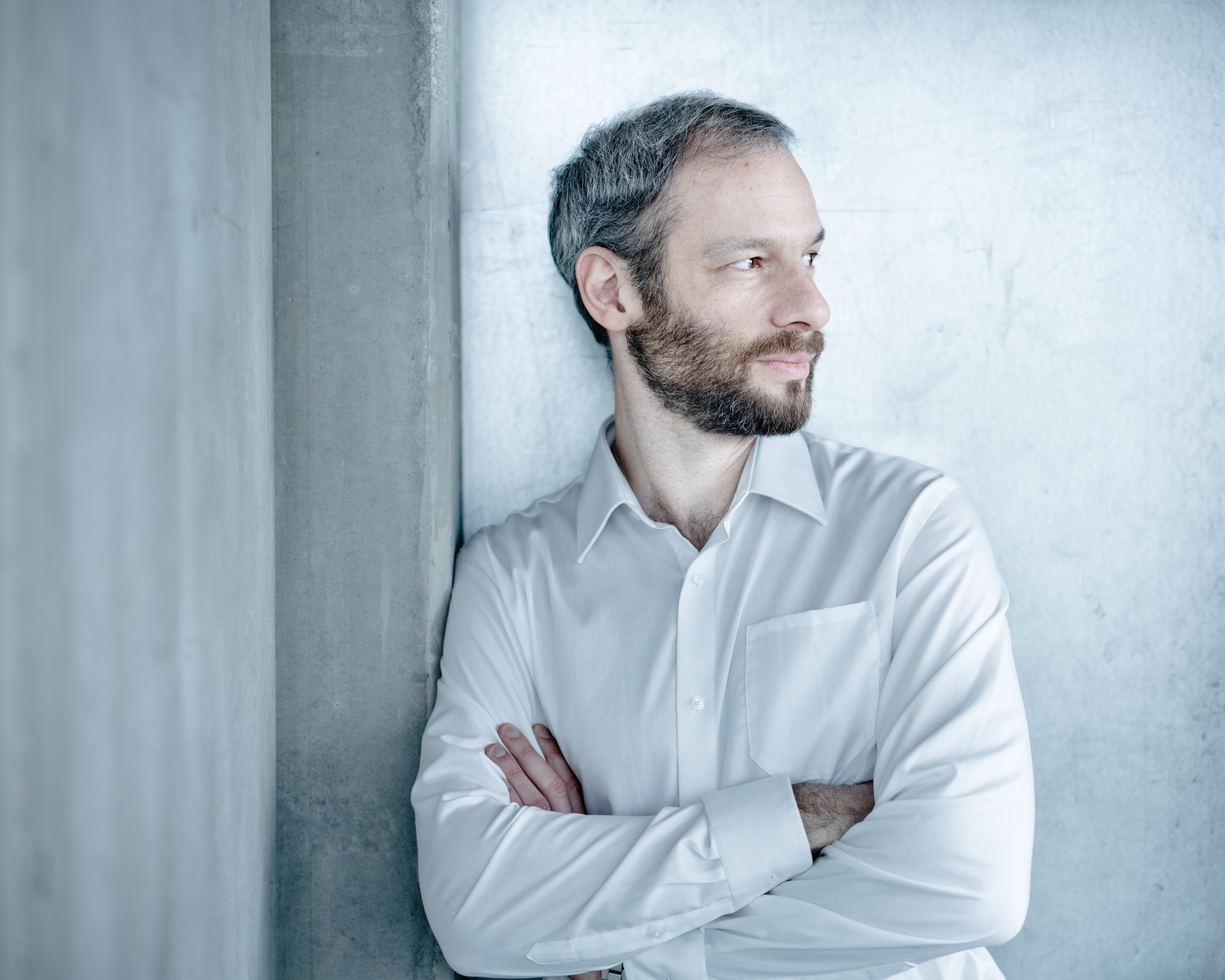 Einer Person mit kurzen grau-schwarzen Haaren und Bart lehnt mit verschränkten Armen an einer Betonwand. Die Person schaut nach rechts und trägt ein weißes Hemd.