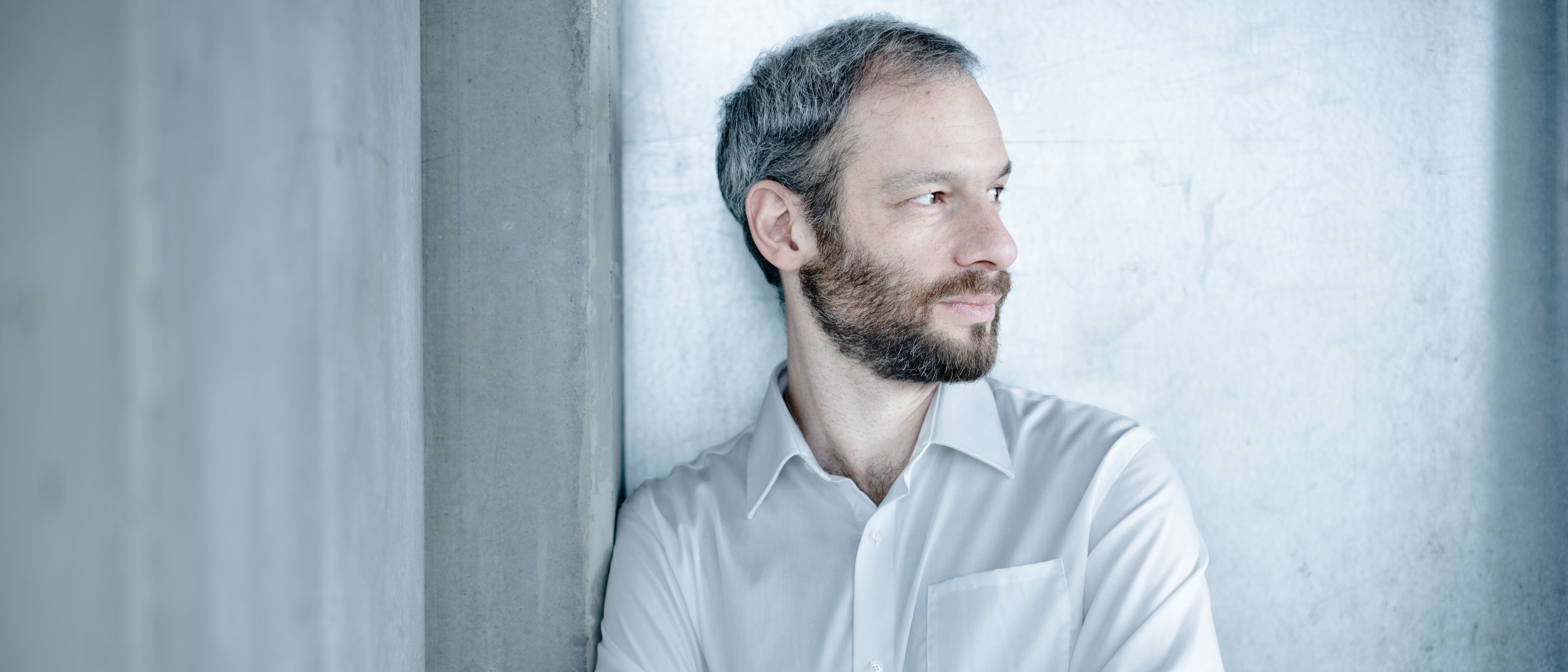 Einer Person mit kurzen grau-schwarzen Haaren und Bart lehnt mit verschränkten Armen an einer Betonwand. Die Person schaut nach rechts und trägt ein weißes Hemd.