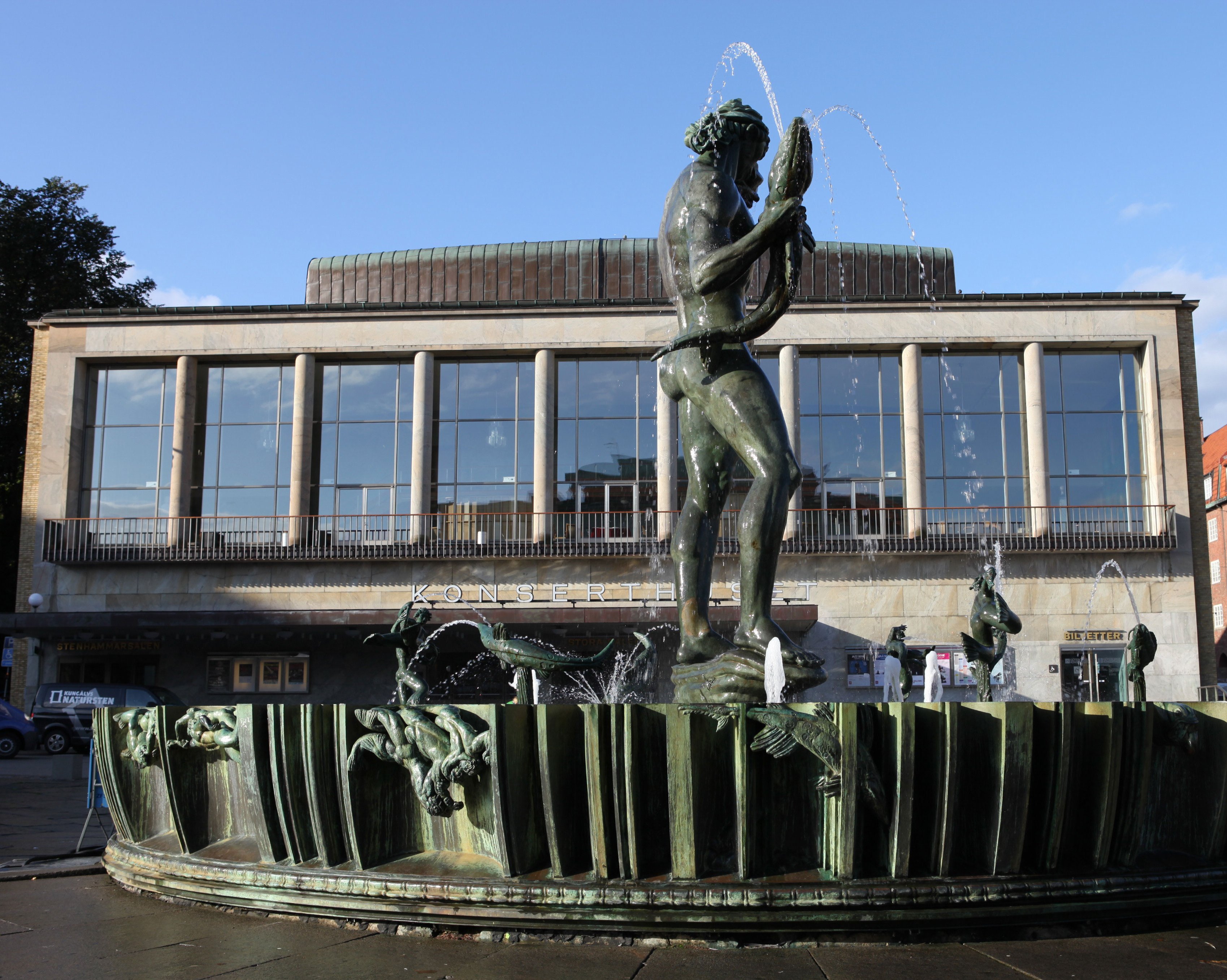 Eine bronzene Neptunstatue steht in einem Brunnen mit einem Fisch in der Hand, der von Wasserstrahlen umspült wird, vor einem modernen Gebäude mit großen Fenstern und ein paar Menschen in der Nähe.
