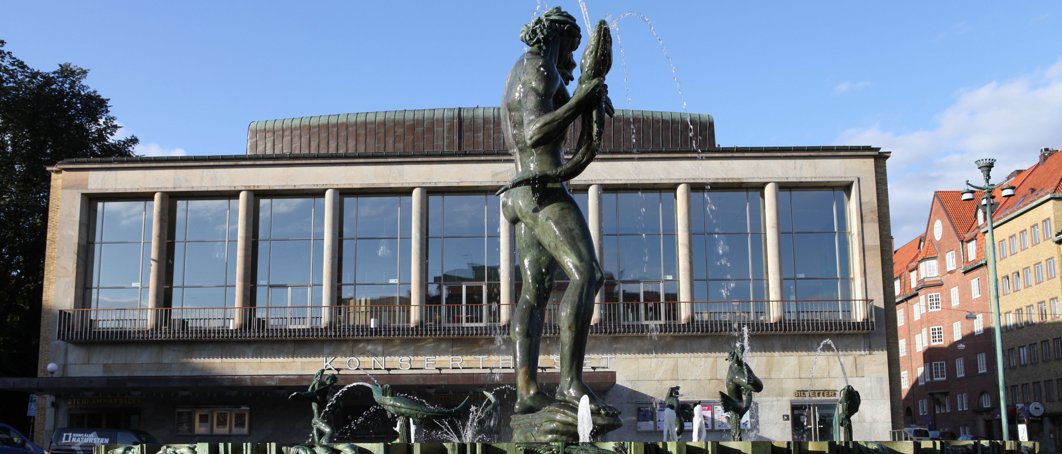 Eine bronzene Neptunstatue steht in einem Brunnen mit einem Fisch in der Hand, der von Wasserstrahlen umspült wird, vor einem modernen Gebäude mit großen Fenstern und ein paar Menschen in der Nähe.