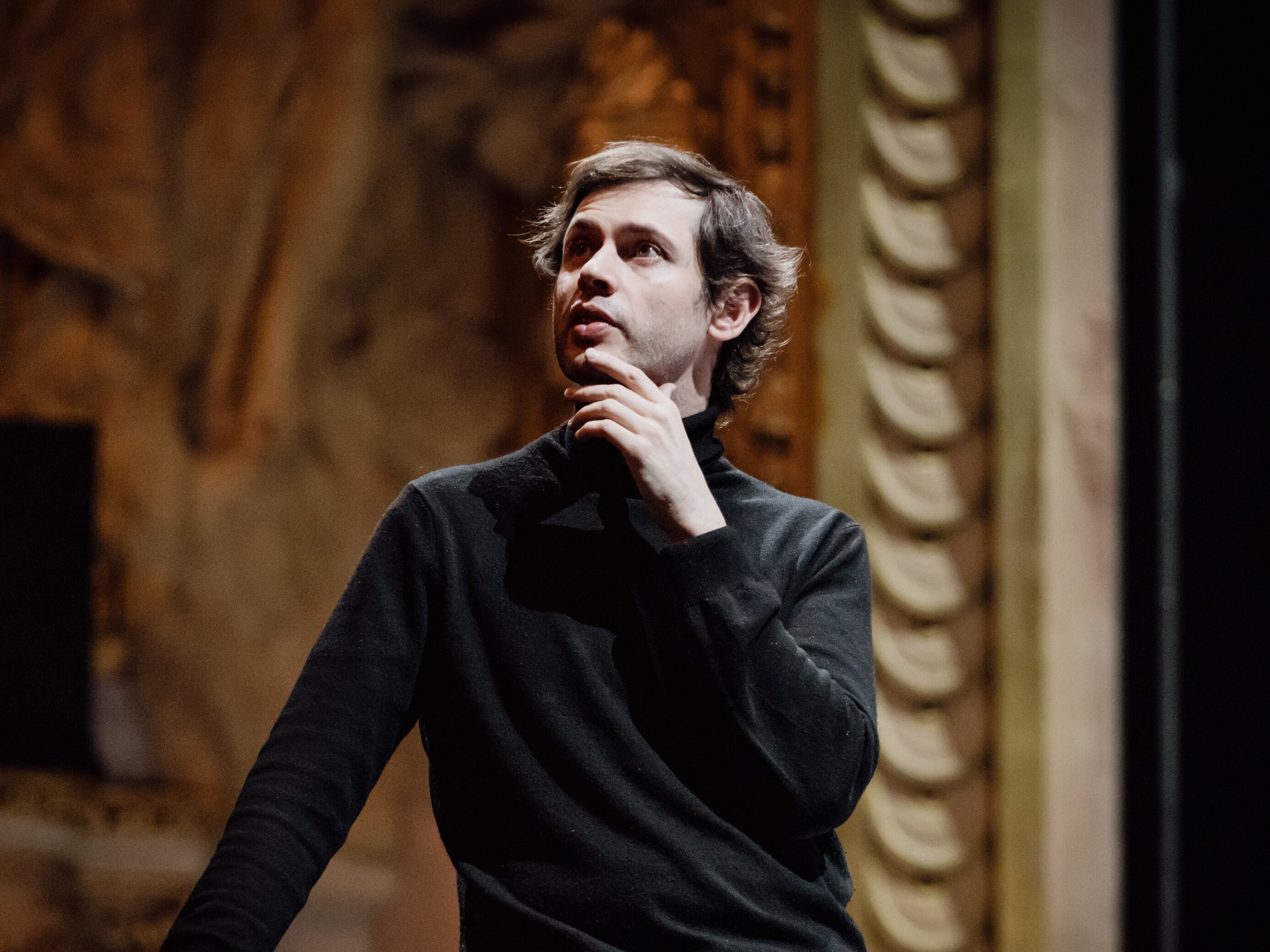 Maxime Pascal in einem schwarzen Pullover steht in einem Haus, stützt sich mit einer Hand auf ein Geländer und berührt nachdenklich sein Kinn, während im Hintergrund kunstvolle architektonische Details zu sehen sind.