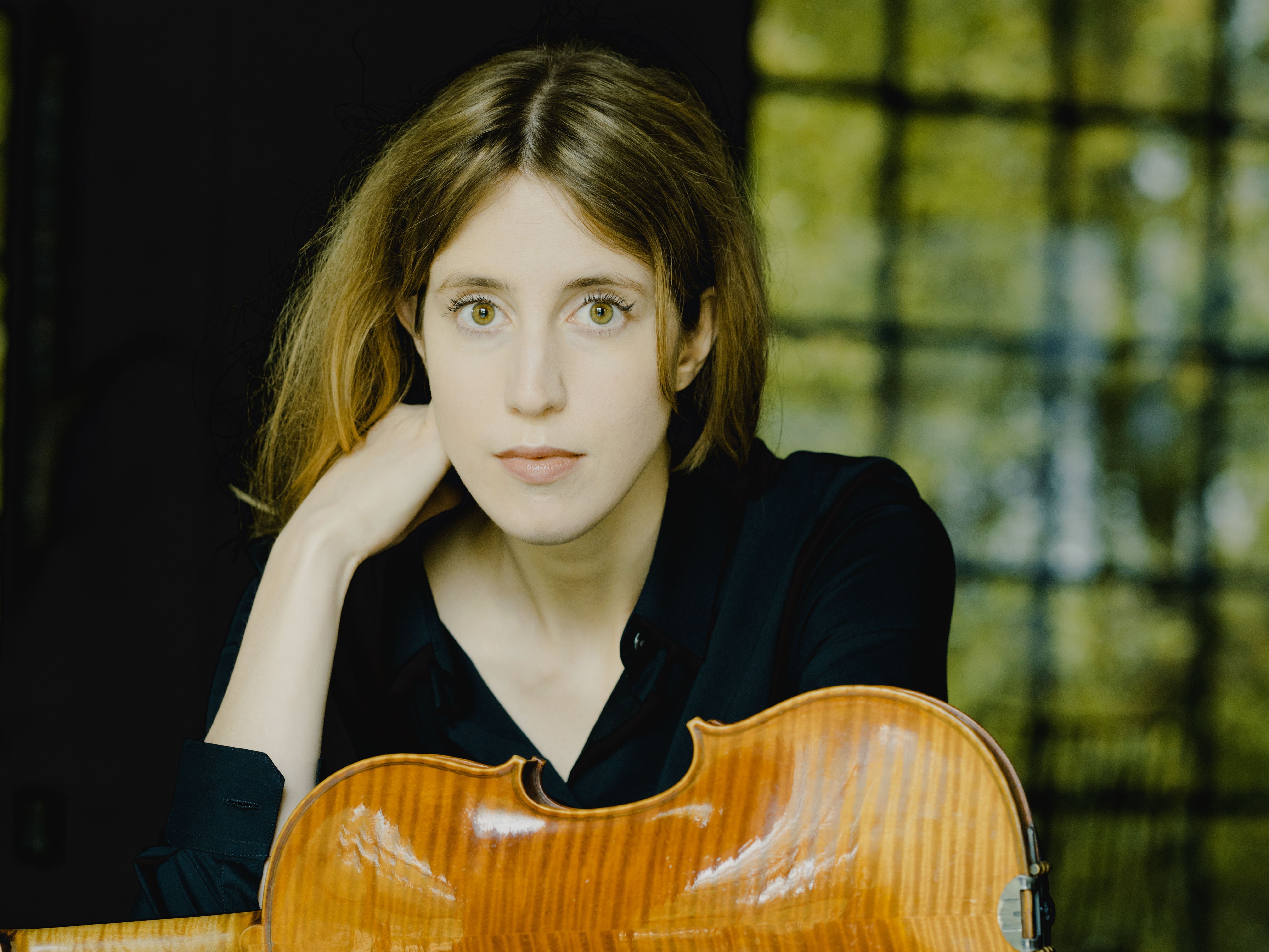 Vilde Frang mit hellbraunem Haar und dunklem Hemd sitzt auf einer spiegelnden Fläche und hat eine Geige vor sich. Sie stützt ihren Kopf auf ihre Hand, und im Hintergrund ist ein Fenster mit Grünpflanzen zu sehen.