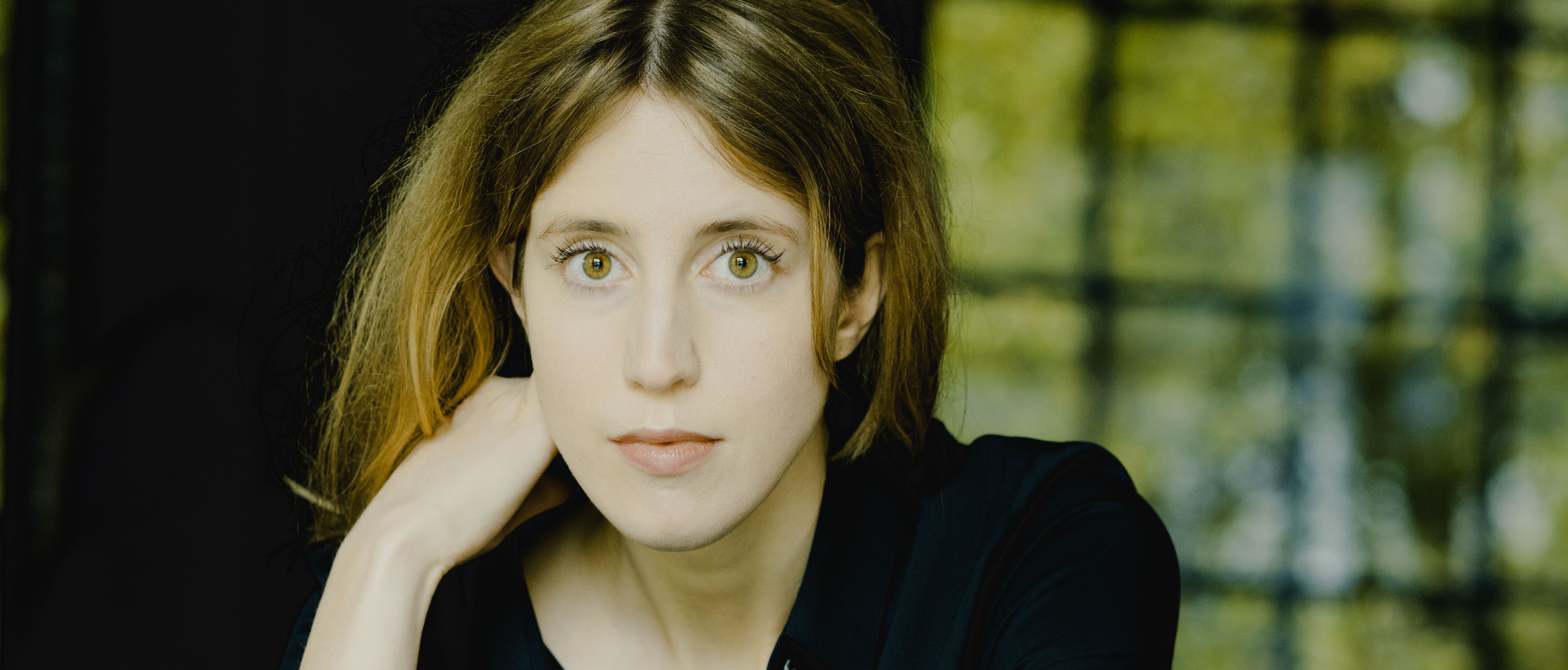 Vilde Frang mit hellbraunem Haar und dunklem Hemd sitzt auf einer spiegelnden Fläche und hat eine Geige vor sich. Sie stützt ihren Kopf auf ihre Hand, und im Hintergrund ist ein Fenster mit Grünpflanzen zu sehen.