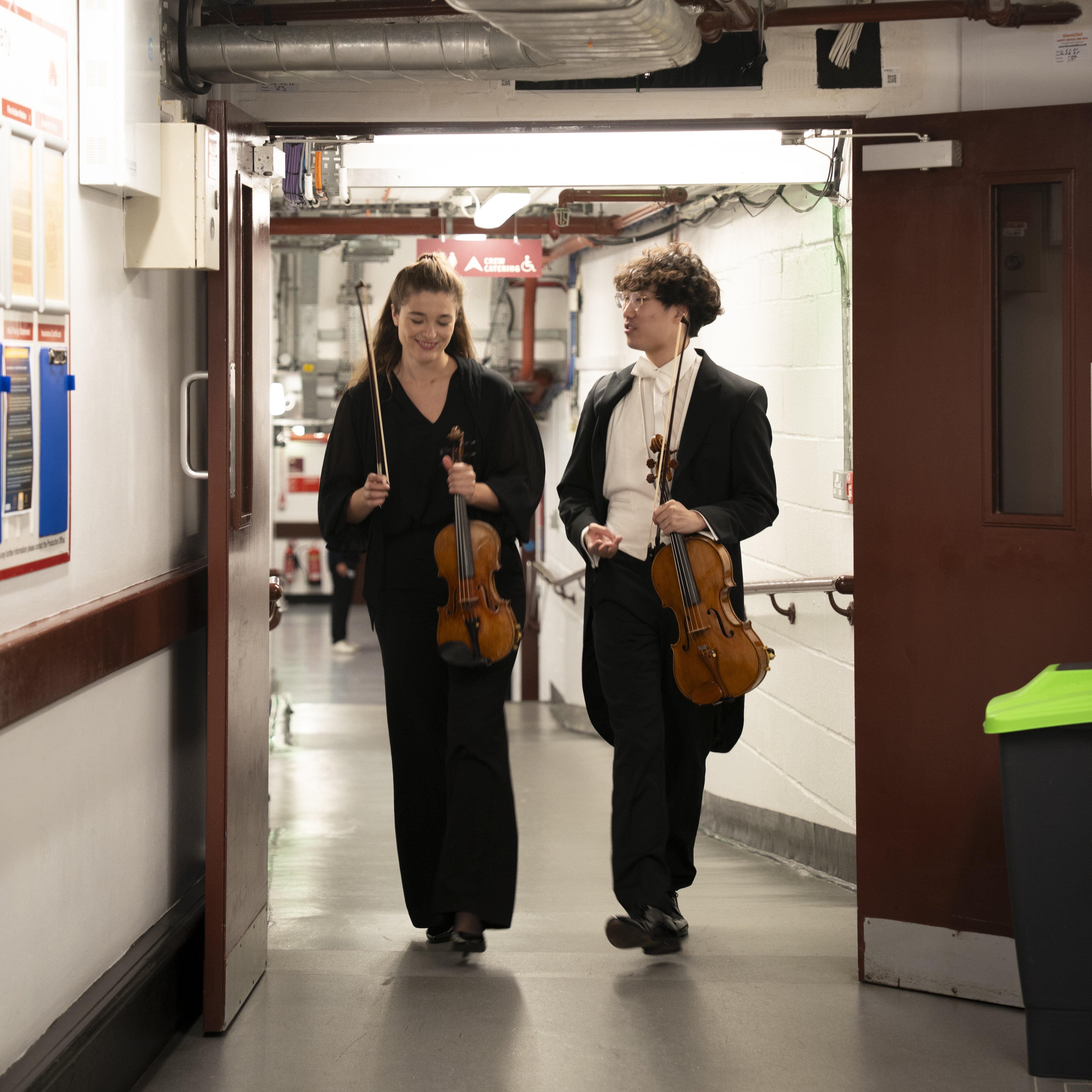 Two musicians in formal black attire walk down a hallway, each holding a violin and smiling at each other. The hallway has exposed pipes and bright lighting overhead.