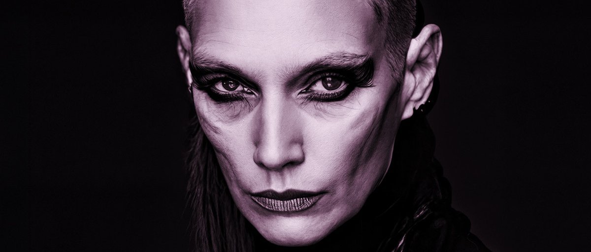 A person with a shaved head and dramatic, dark makeup stares intensely at the camera. Their sharp cheek contour and bold eyeliner create a striking, high-fashion look against a dark background.
