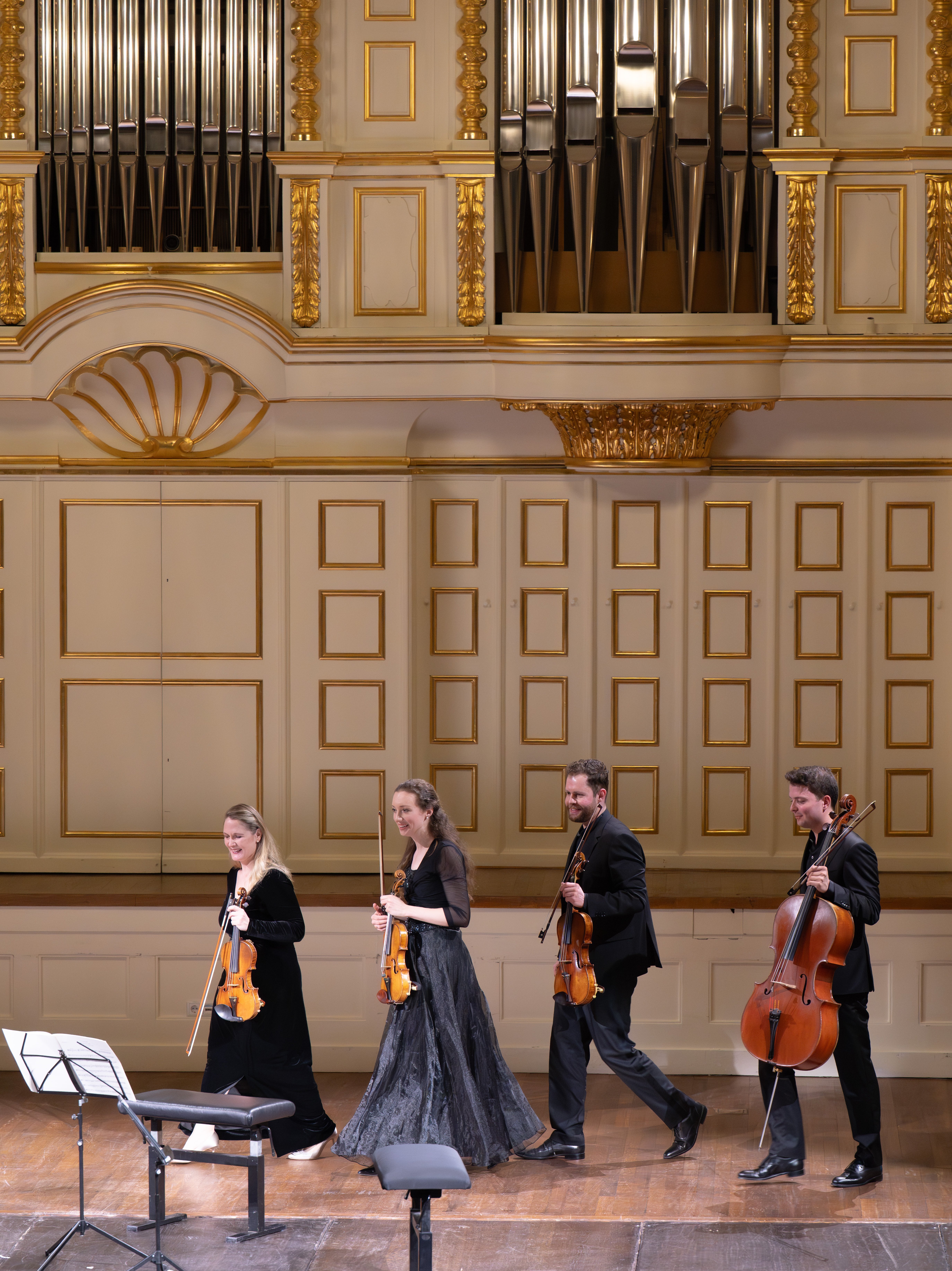 Vier Musiker in förmlicher Kleidung und mit Streichinstrumenten in der Hand betreten die Bühne eines großen Konzertsaals mit kunstvollen goldenen Verzierungen und einer Pfeifenorgel im Hintergrund.