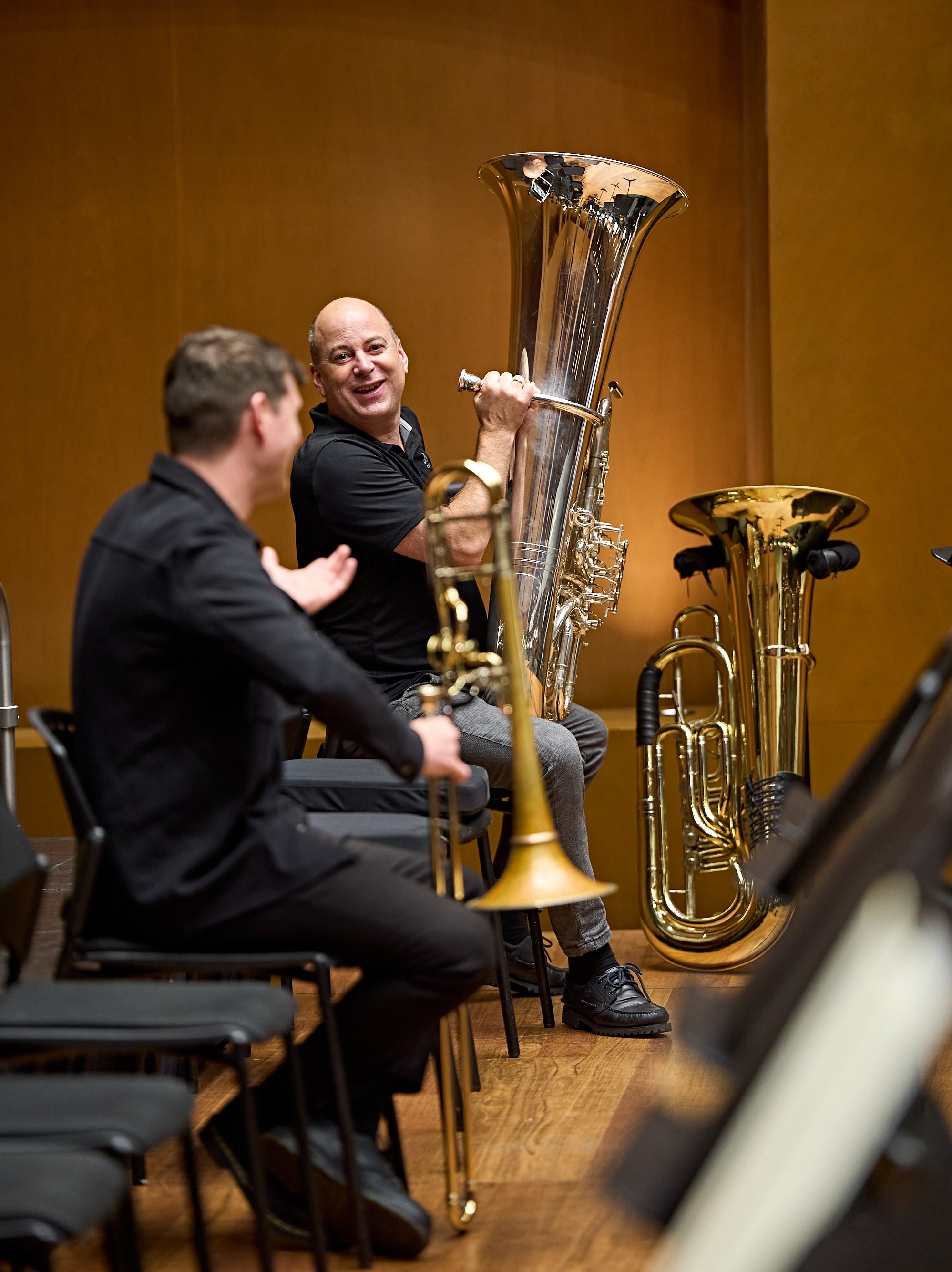 Ein lächelnder Musiker sitzt mit einer silbernen Tuba auf der Bühne, während im Vordergrund ein Posaunist sitzt.