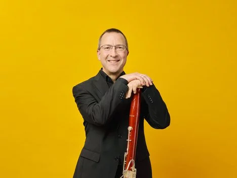 A smiling Markus Weidmann wearing glasses and a black suit stands against a yellow background, resting his hands on a bassoon.