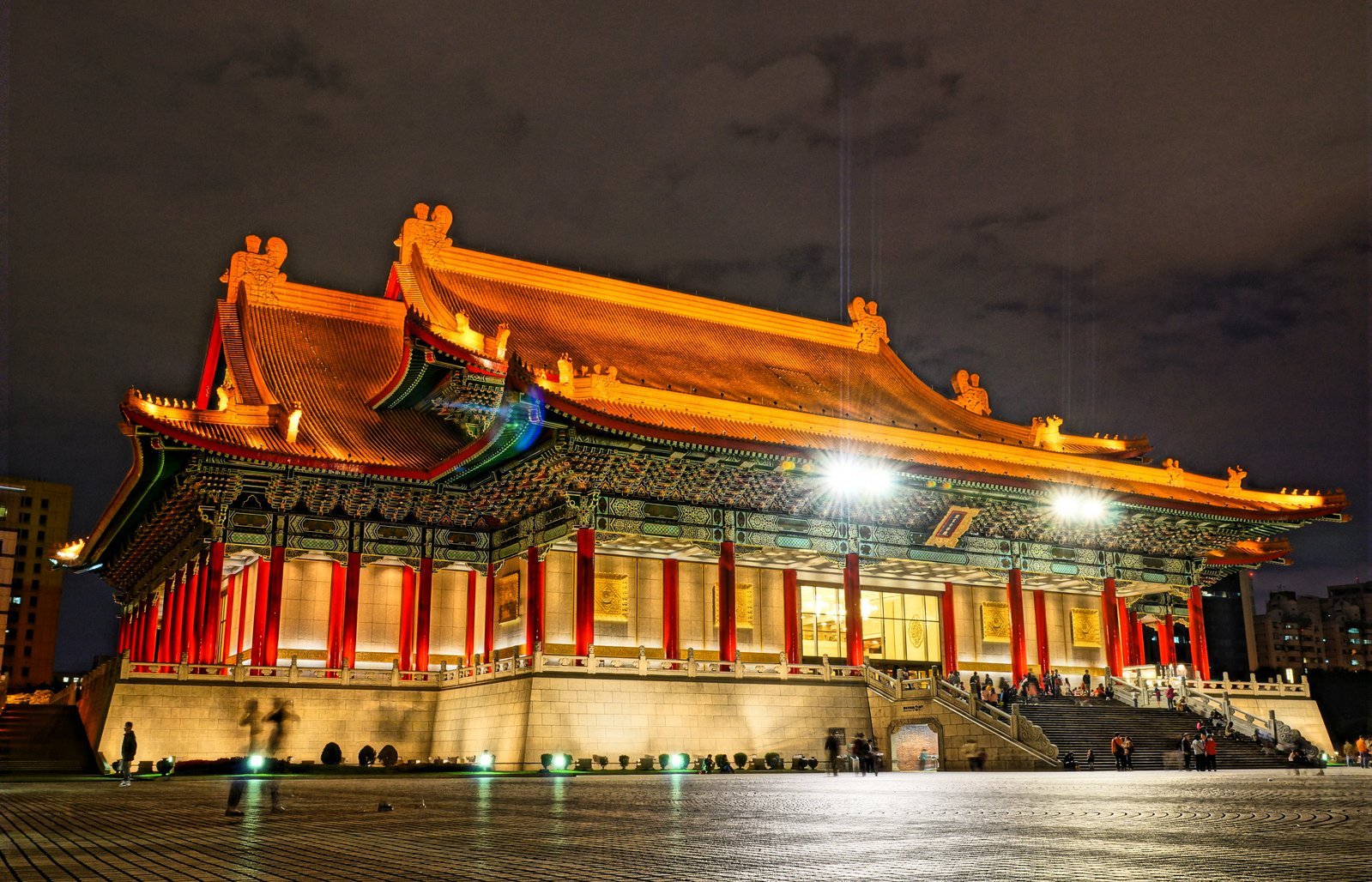 A large traditional Chinese building with ornate, illuminated roofs and red columns stands brightly lit at night, with a spacious stone plaza and people gathered near the entrance.