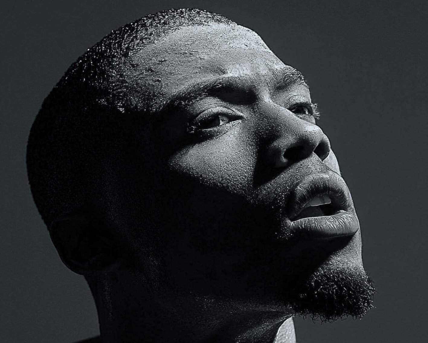 A black and white close-up portrait of a man looking upwards. The dramatic lighting casts deep shadows across his face, highlighting his textured skin and contemplative expression.