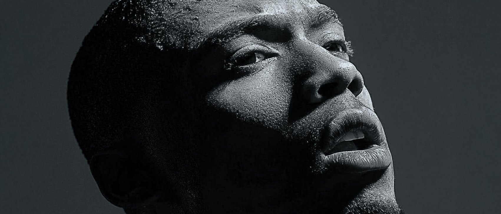 A black and white close-up portrait of a man looking upwards. The dramatic lighting casts deep shadows across his face, highlighting his textured skin and contemplative expression.