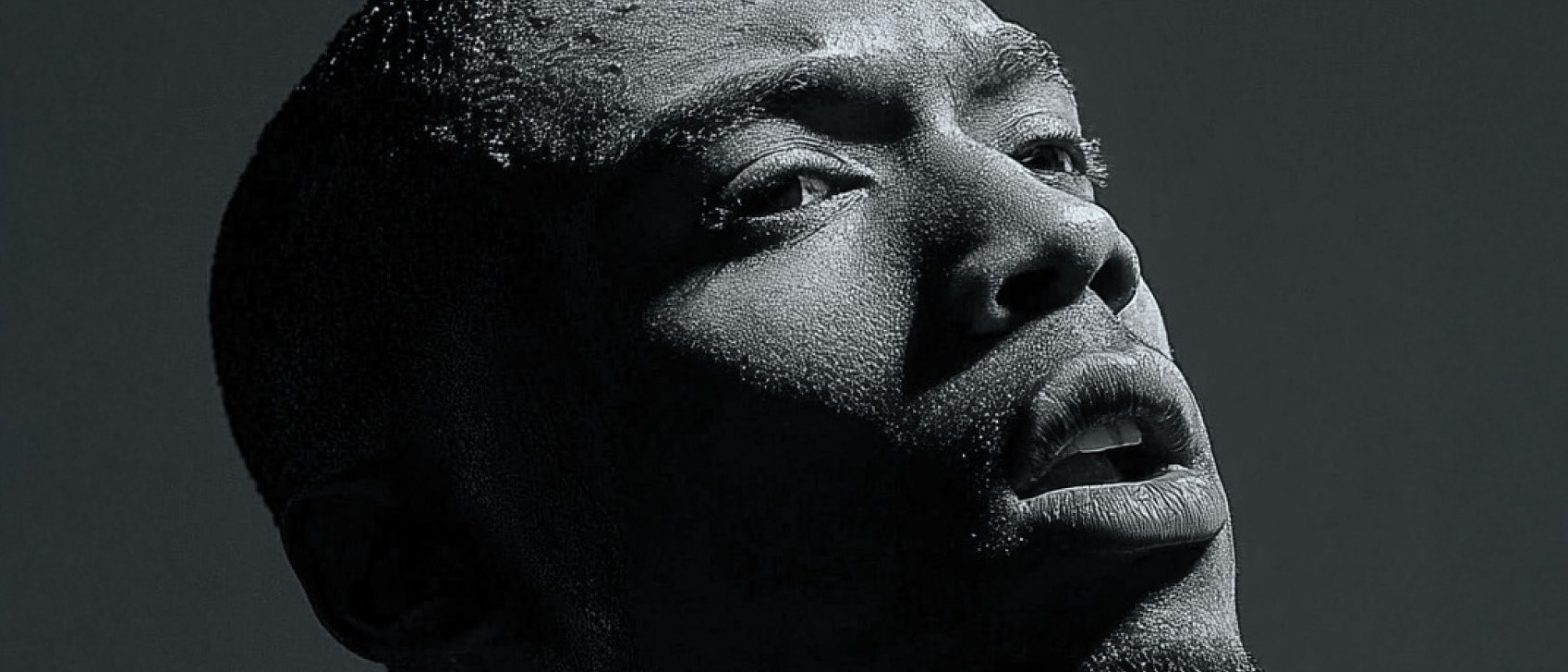 Ein Schwarz-Weiß-Porträt eines Mannes in Nahaufnahme, der nach oben blickt. Die dramatische Beleuchtung wirft starke Schatten auf sein Gesicht und hebt seine strukturierte Haut und seinen nachdenklichen Ausdruck hervor.