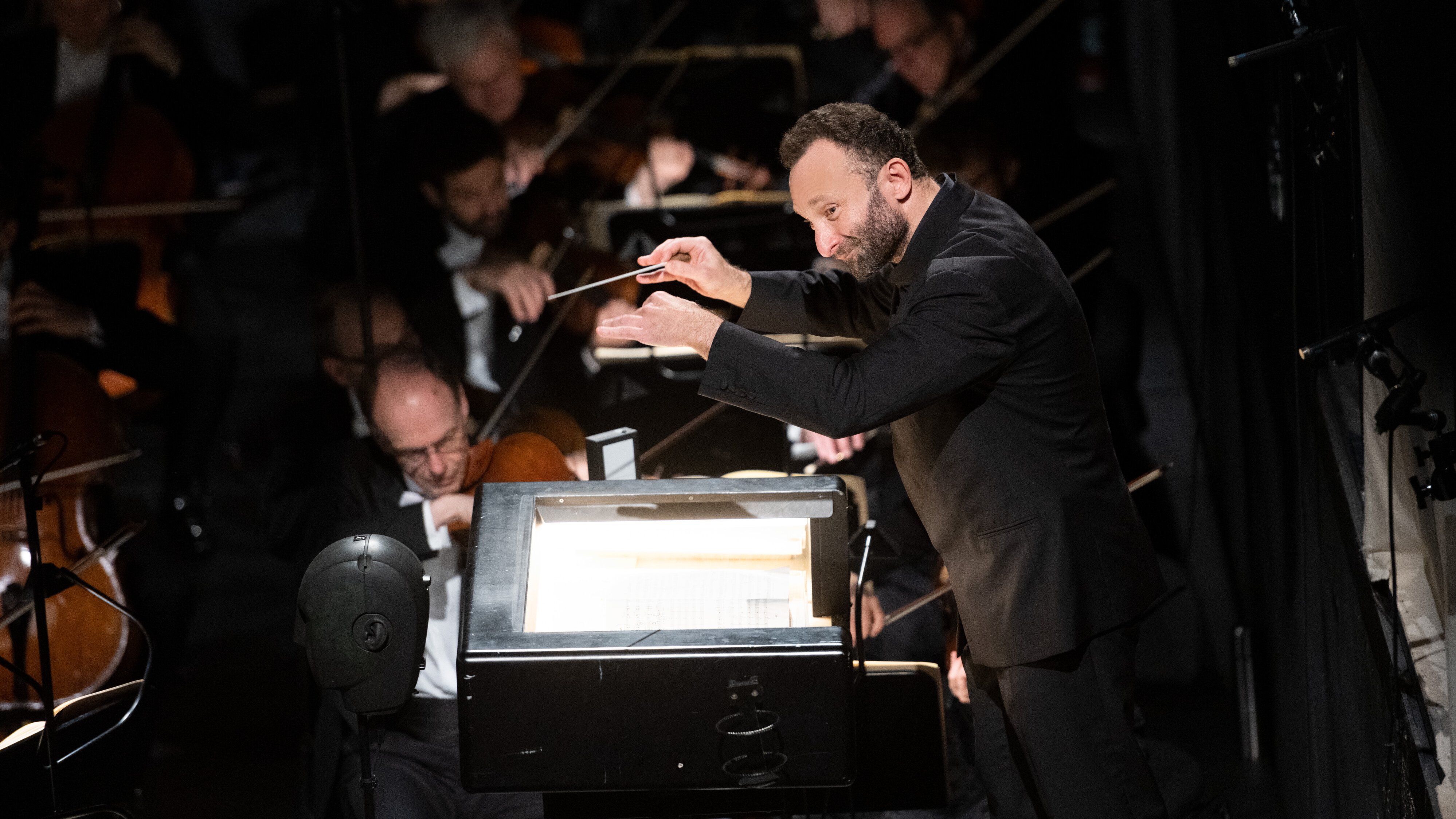 Kirill Petrenko im schwarzen Anzug leitet energisch ein Orchester mit erhobenem Taktstock, während im Hintergrund bei gedämpfter Beleuchtung Musiker auf Streichinstrumenten spielen.