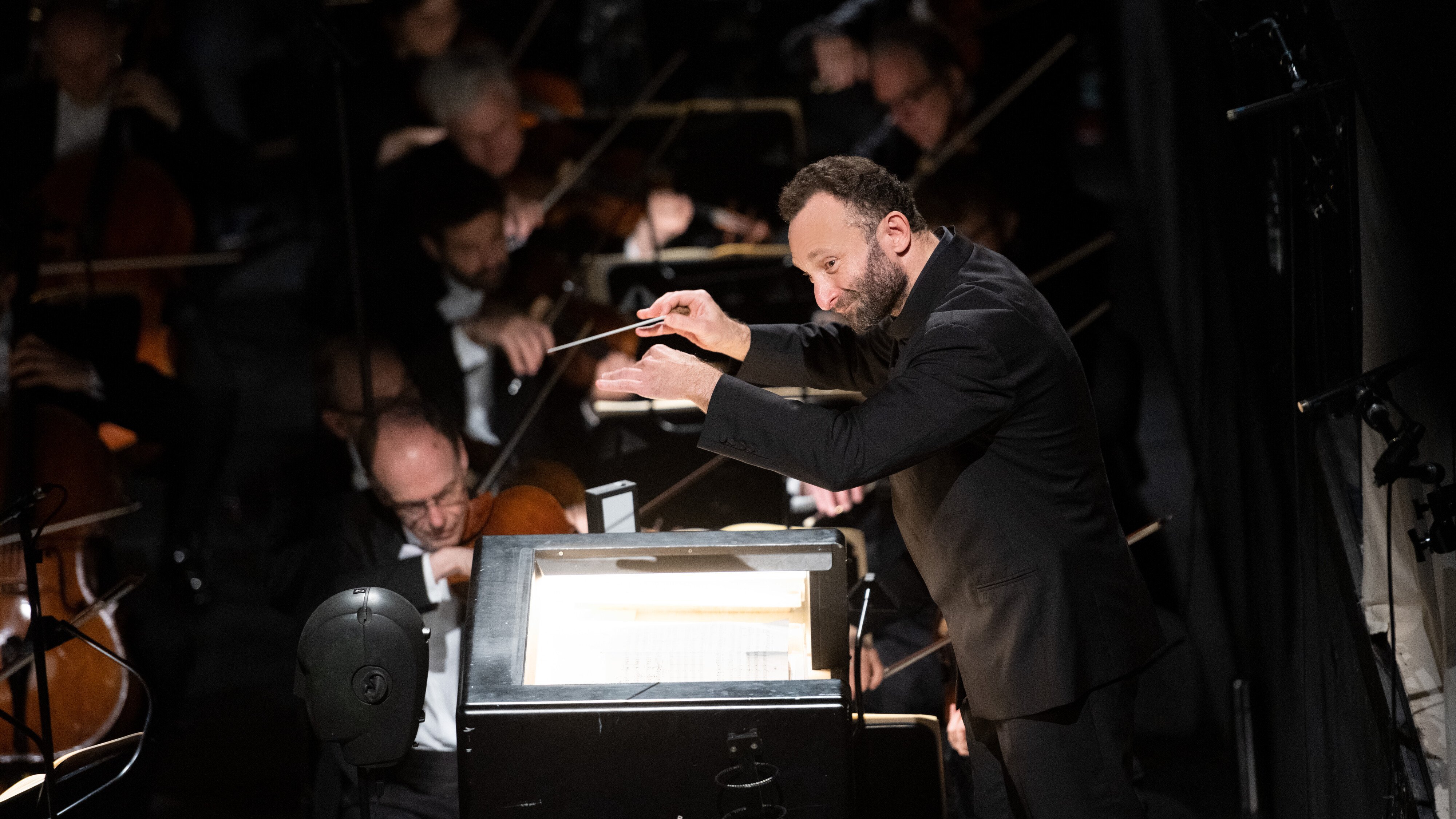 Kirill Petrenko im schwarzen Anzug leitet energisch ein Orchester mit erhobenem Taktstock, während im Hintergrund bei gedämpfter Beleuchtung Musiker auf Streichinstrumenten spielen.