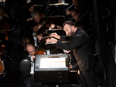 Kirill Petrenko im schwarzen Anzug leitet energisch ein Orchester mit erhobenem Taktstock, während im Hintergrund bei gedämpfter Beleuchtung Musiker auf Streichinstrumenten spielen.