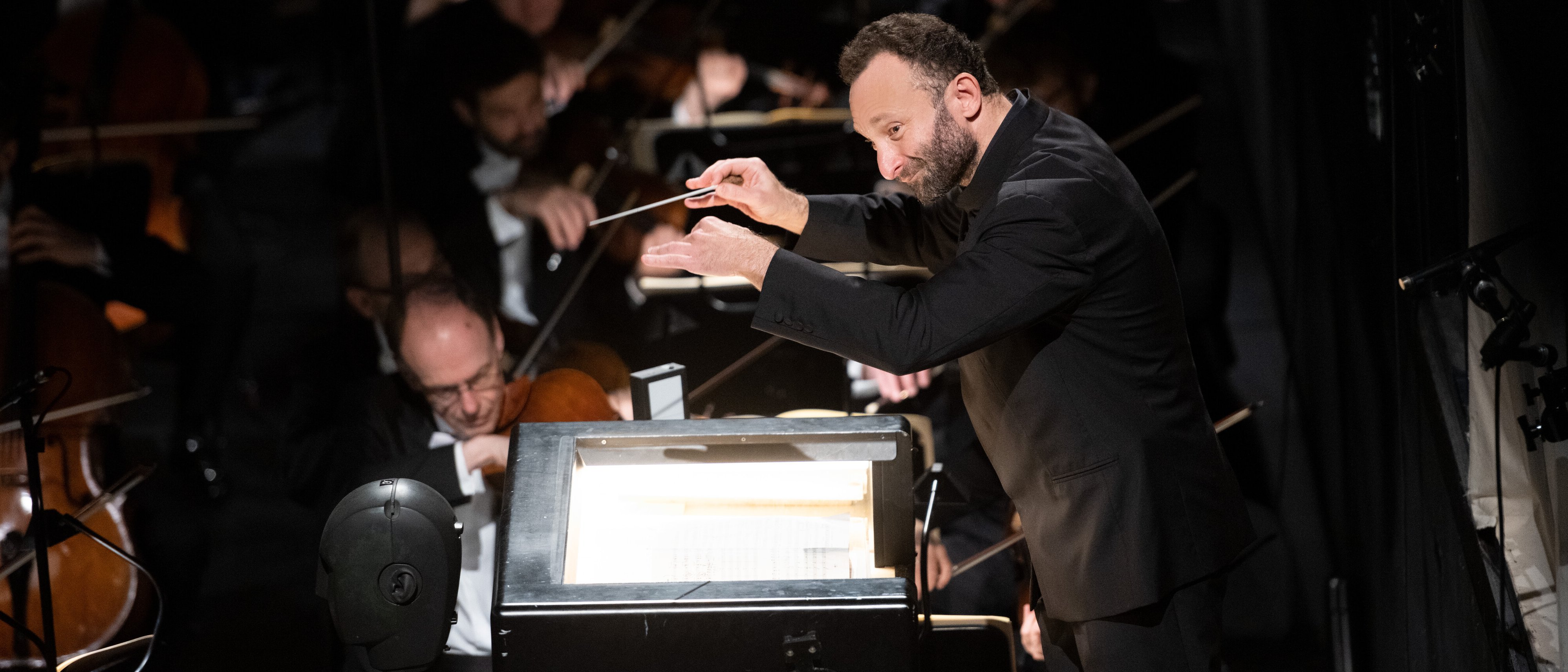 Kirill Petrenko im schwarzen Anzug leitet energisch ein Orchester mit erhobenem Taktstock, während im Hintergrund bei gedämpfter Beleuchtung Musiker auf Streichinstrumenten spielen.