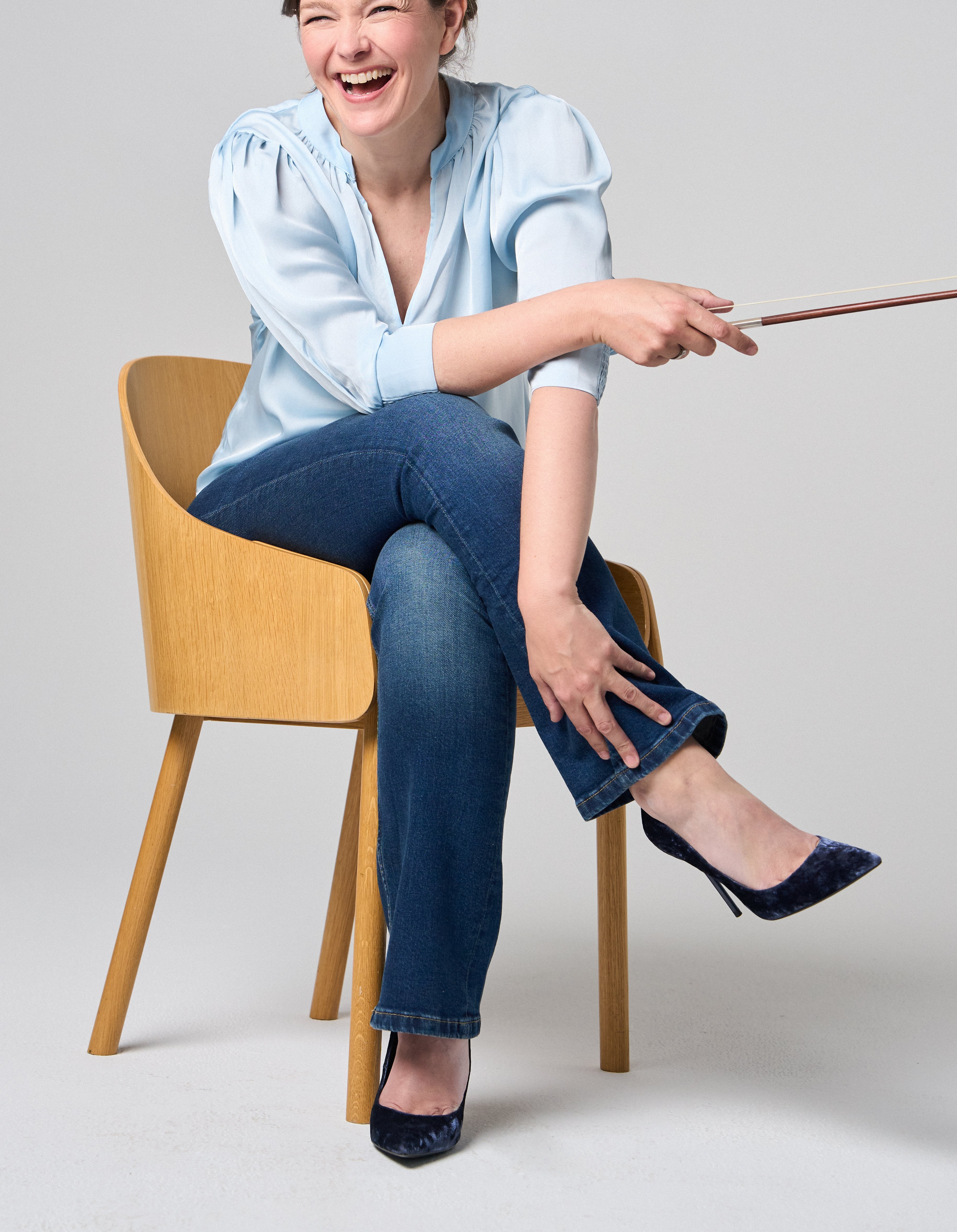 Janine Jansen sitzt auf einem Stuhl, eine Violine in den Händen. Sie trägt Jeans und eine hellblaue Bluse und blickt lächelnd in die Kamera.