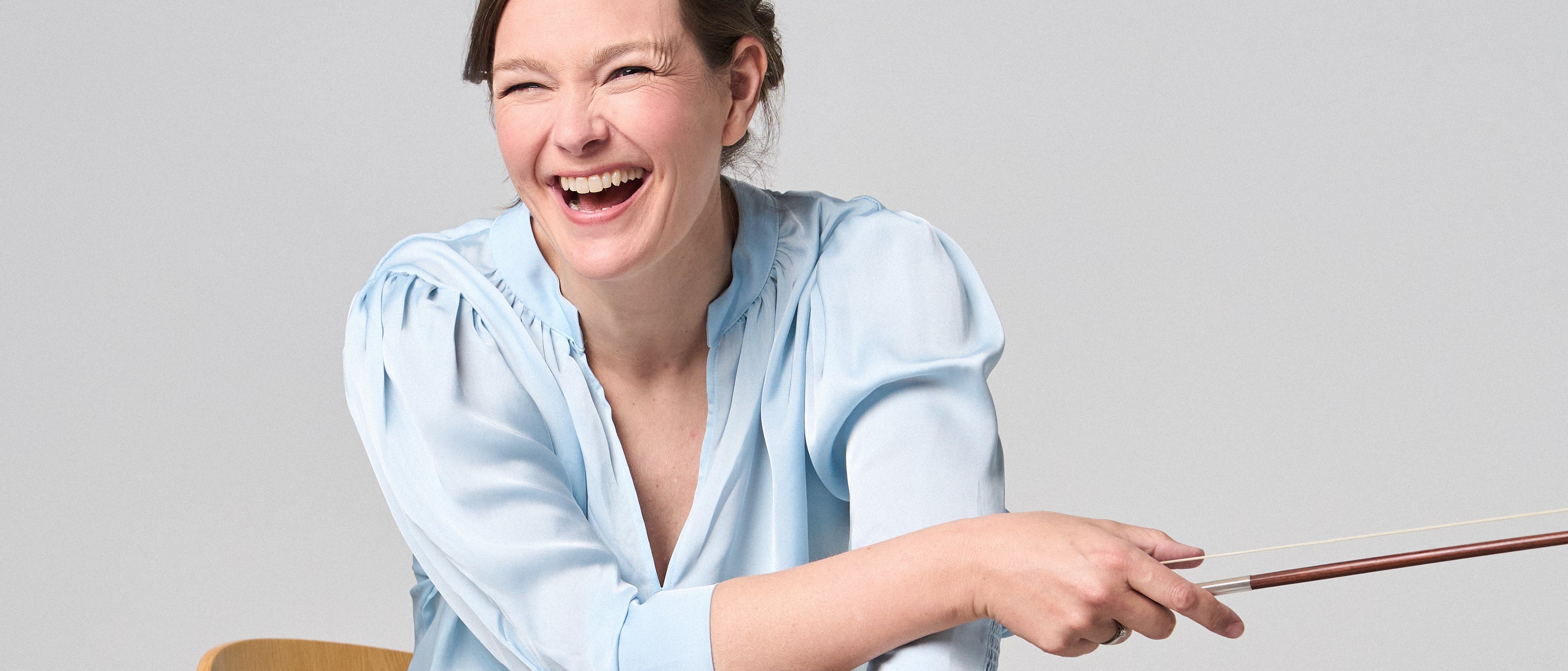 Janine Jansen is sitting on a chair, a violin in her hands. She is wearing jeans and a light blue blouse and looks into the camera with a smile.