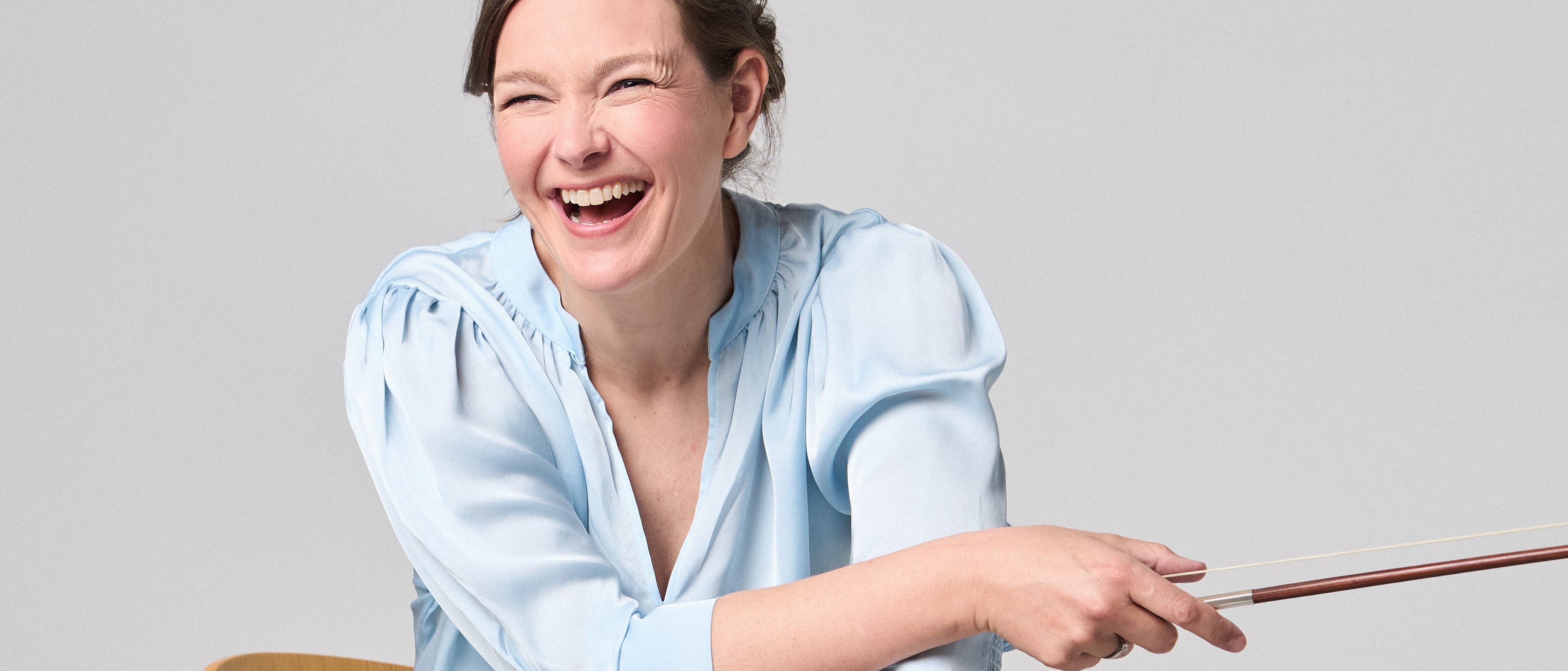 Janine Jansen is sitting on a chair, a violin in her hands. She is wearing jeans and a light blue blouse and looks into the camera with a smile.