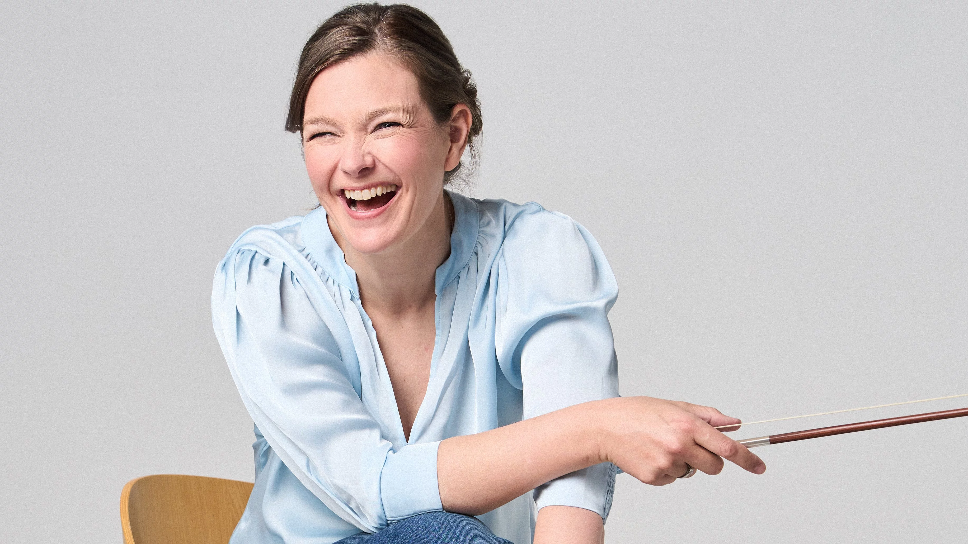 Janine Jansen is sitting on a chair, a violin in her hands. She is wearing jeans and a light blue blouse and looks into the camera with a smile.