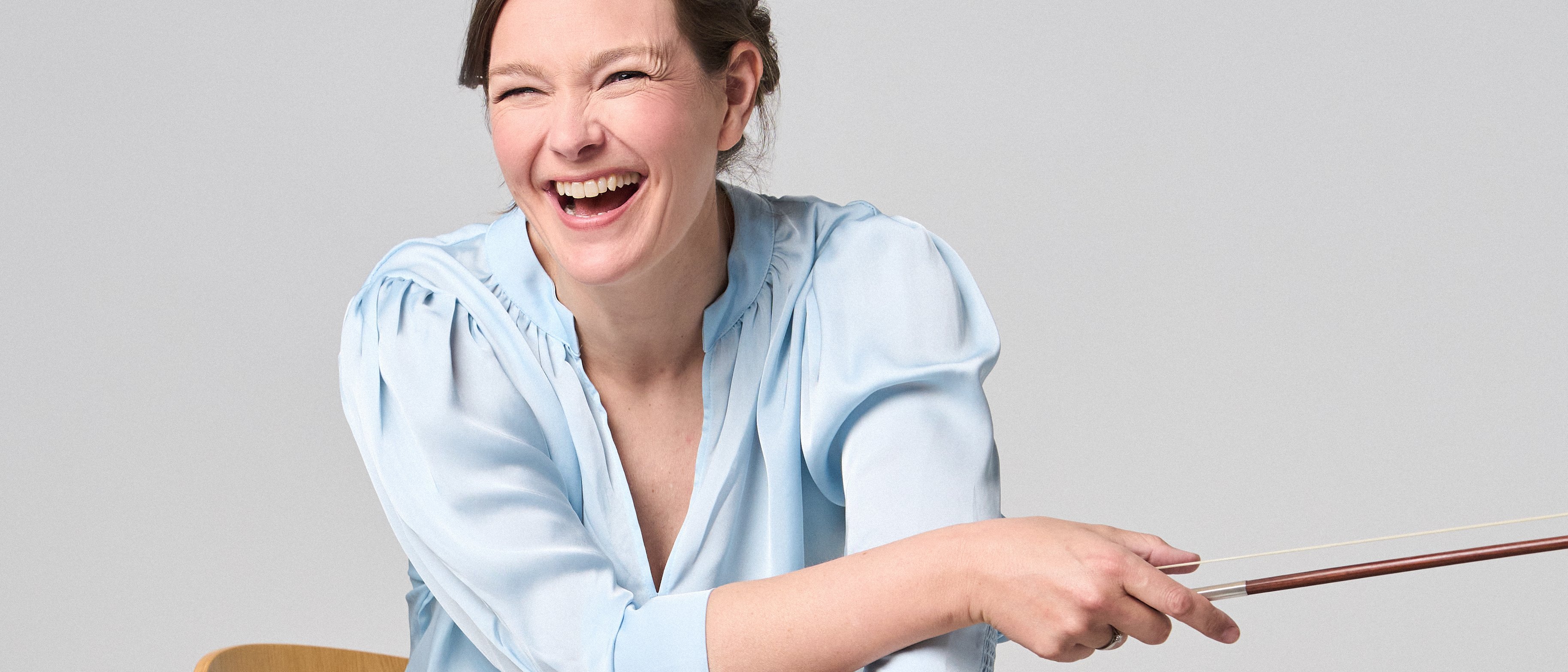 Janine Jansen is sitting on a chair, a violin in her hands. She is wearing jeans and a light blue blouse and looks into the camera with a smile.