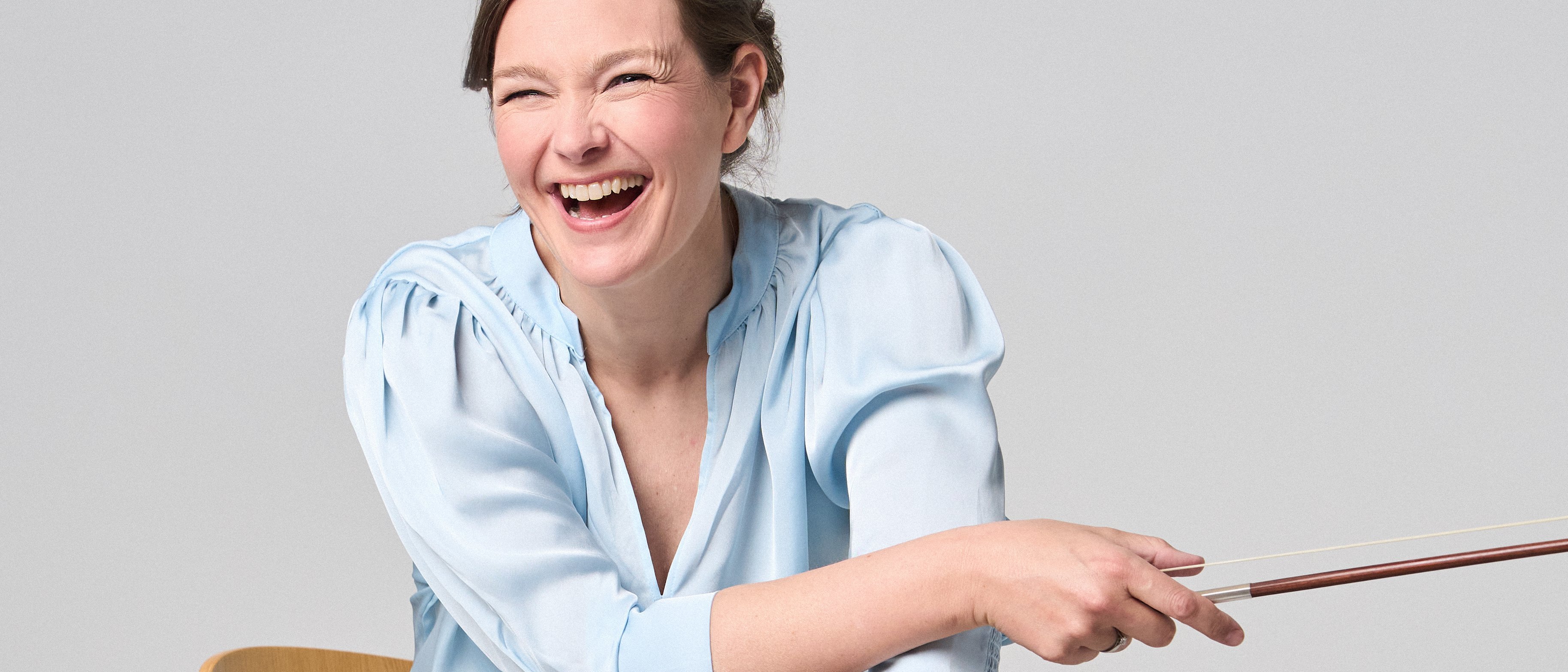 Janine Jansen is sitting on a chair, a violin in her hands. She is wearing jeans and a light blue blouse and looks into the camera with a smile.