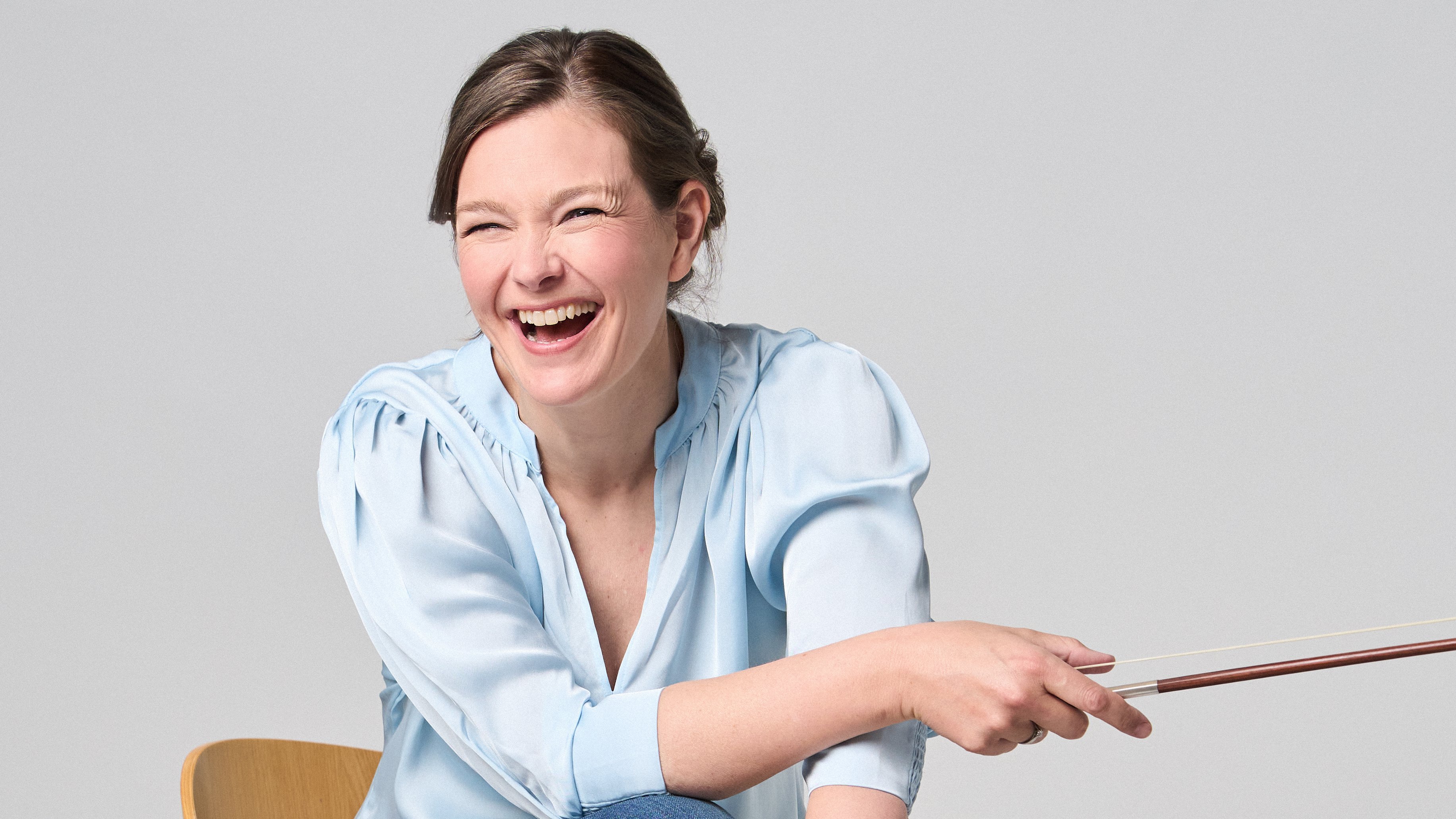 Janine Jansen sitzt auf einem Stuhl, eine Violine in den Händen. Sie trägt Jeans und eine hellblaue Bluse und blickt lächelnd in die Kamera.