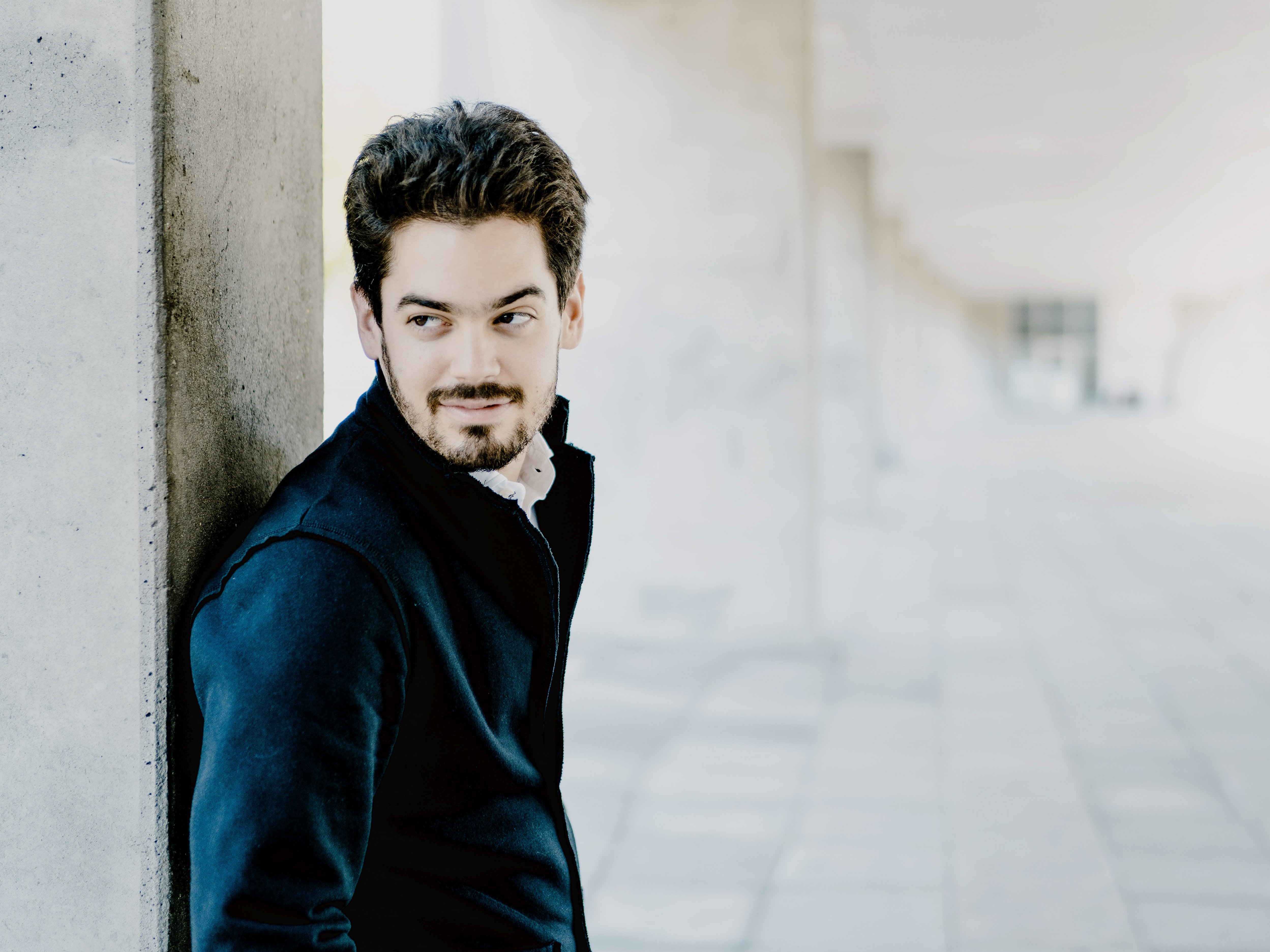 Das Bild zeigt Lahav Shani mit Bart und dunklem Haar, der in einem Flur an einem Betonpfeiler lehnt. Er trägt eine dunkle Jacke und Hose und blickt nachdenklich zur Seite. Die Szenerie umfasst einen gefliesten Boden und eine Decke mit modernen Designelementen.