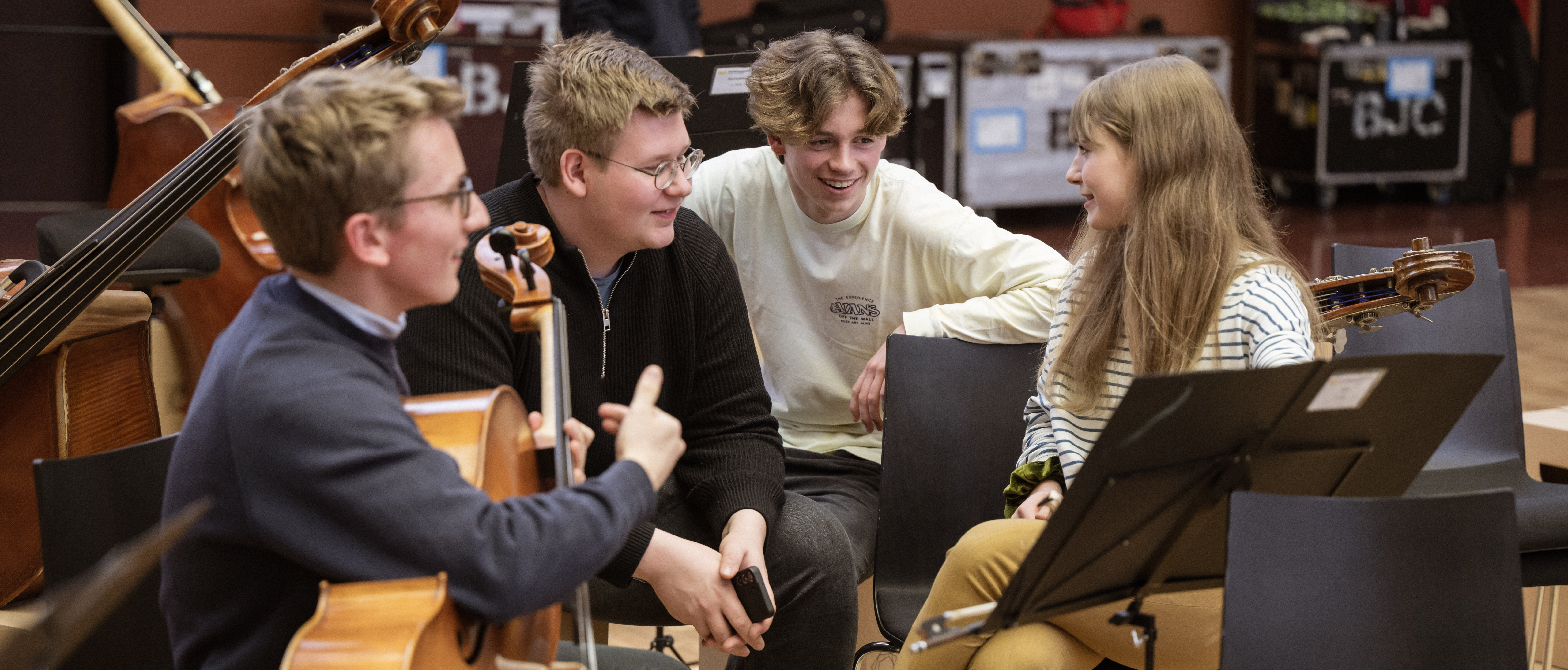 Vier junge Musiker sitzen in einem Proberaum zusammen, lächeln und unterhalten sich. Einer hält ein Cello, ein anderer ein Telefon, und um sie herum sind ein Notenständer und andere Instrumente zu sehen.