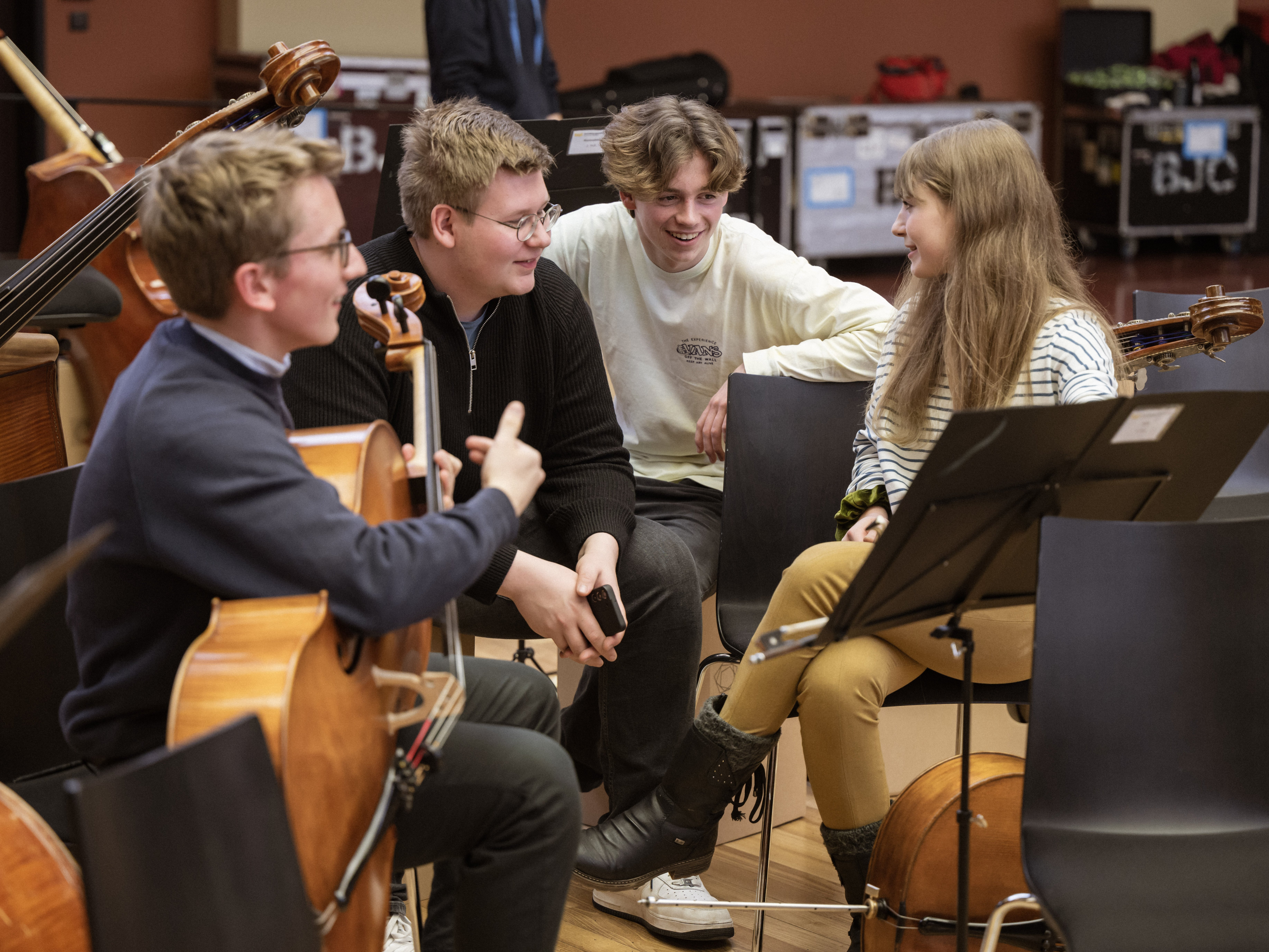 Vier junge Musiker sitzen in einem Proberaum zusammen, lächeln und unterhalten sich. Einer hält ein Cello, ein anderer ein Telefon, und um sie herum sind ein Notenständer und andere Instrumente zu sehen.
