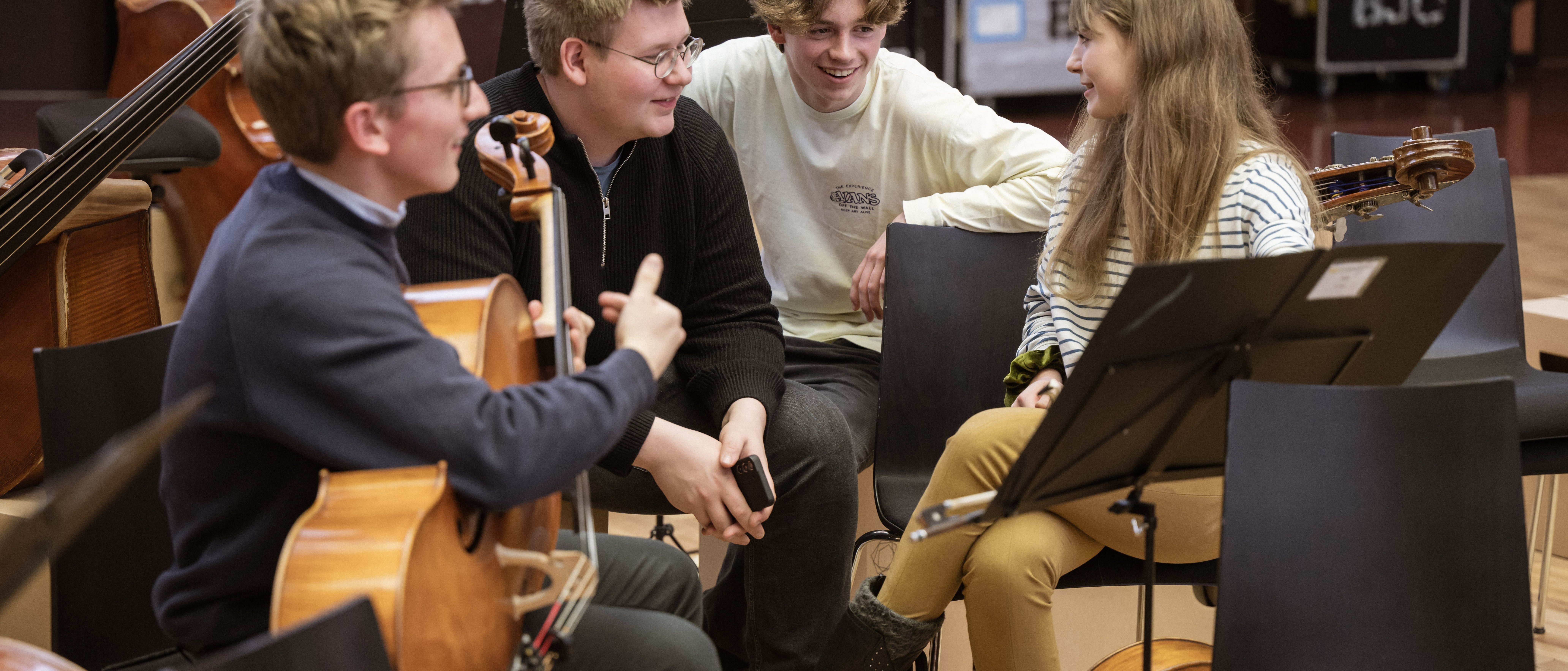 Vier junge Musiker sitzen in einem Proberaum zusammen, lächeln und unterhalten sich. Einer hält ein Cello, ein anderer ein Telefon, und um sie herum sind ein Notenständer und andere Instrumente zu sehen.