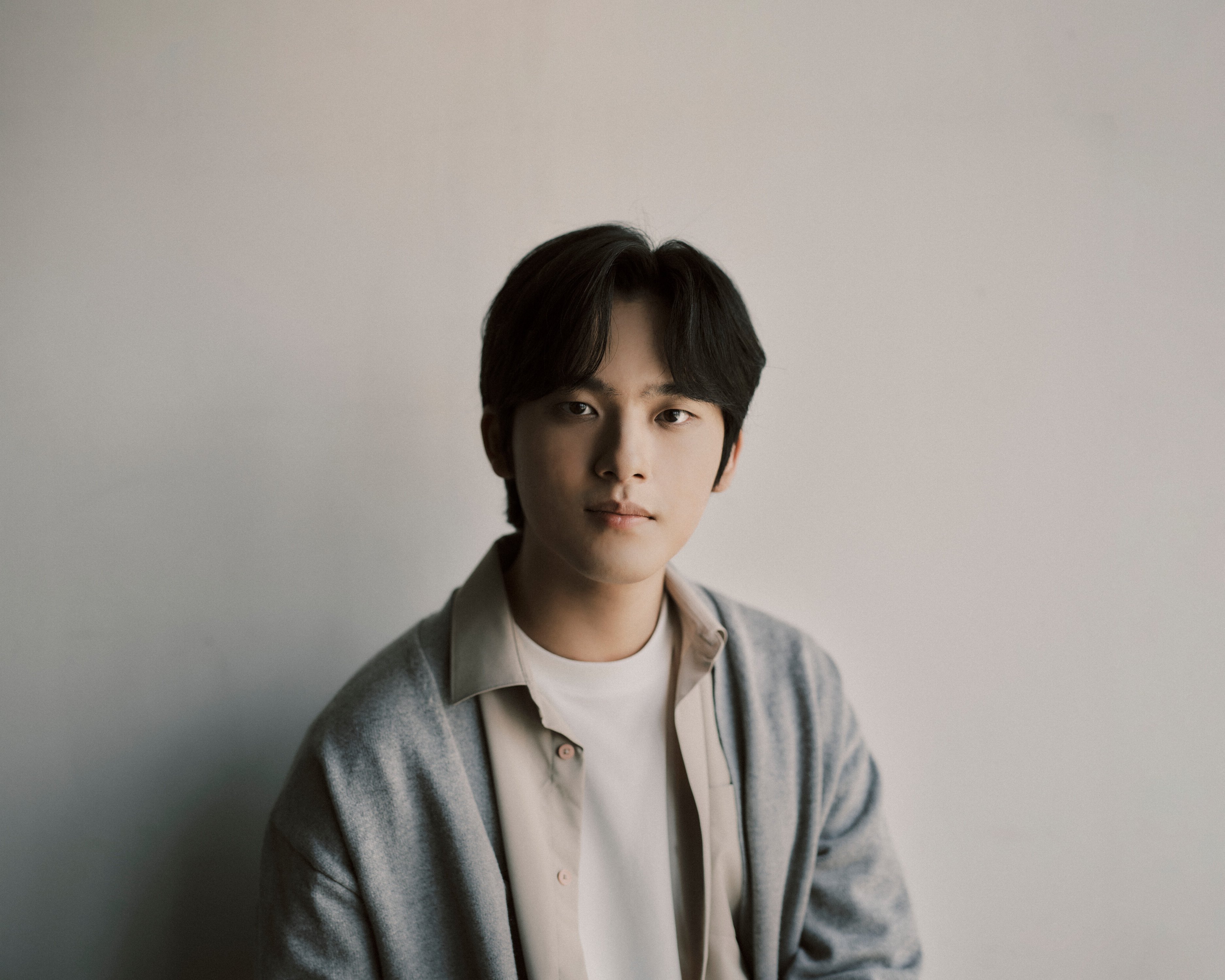 Yunchan Lim mit mittellangen dunklen Haaren, die ein weißes Hemd und eine graue Strickjacke trägt, sitzt vor einem schlichten hellen Hintergrund und schaut ruhig in die Kamera.