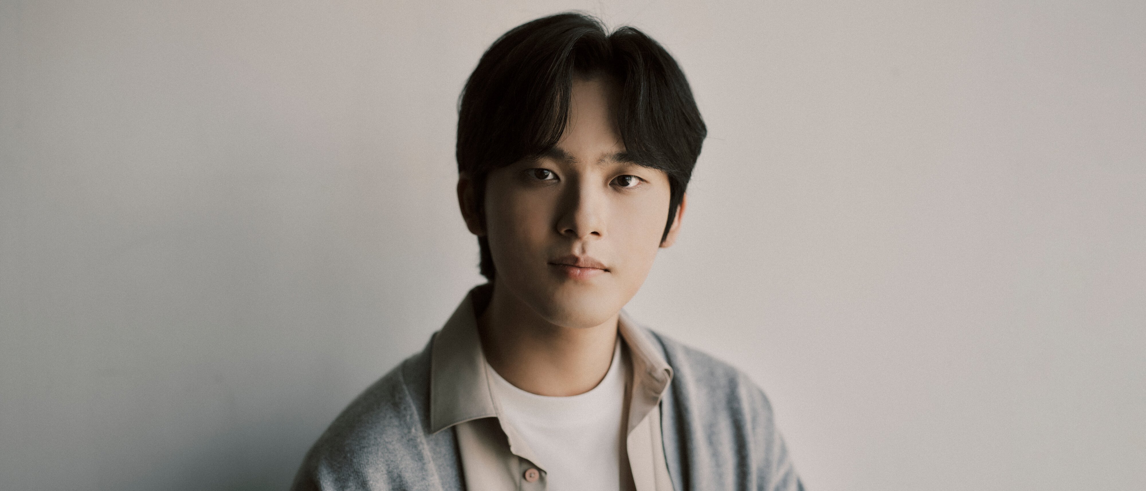 Yunchan Lim mit mittellangen dunklen Haaren, die ein weißes Hemd und eine graue Strickjacke trägt, sitzt vor einem schlichten hellen Hintergrund und schaut ruhig in die Kamera.